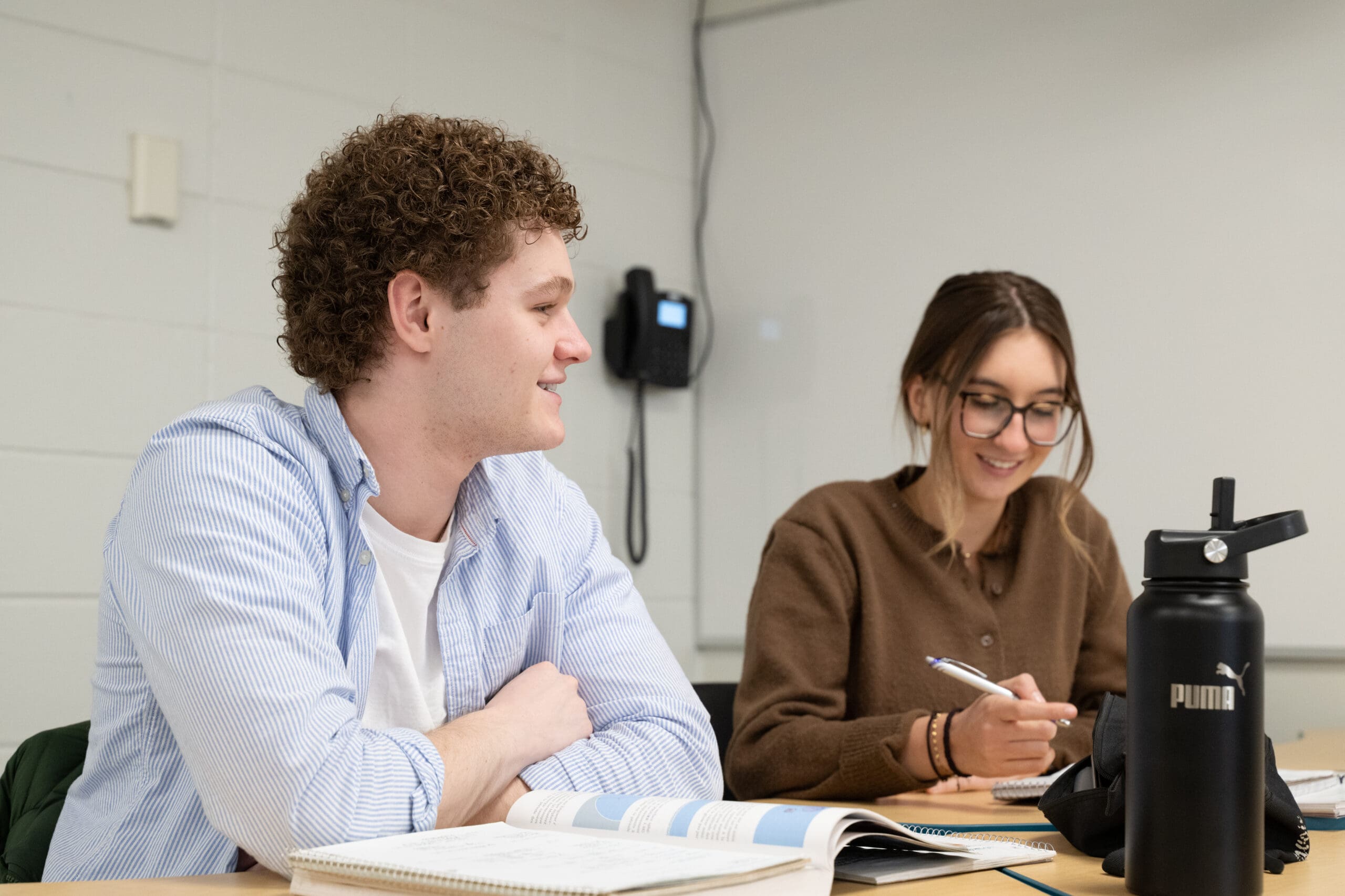 Deux élèves sont assis à une table dans une salle de classe ; ils sourient et prennent des notes dans leurs cahiers. L'un a les cheveux bouclés et porte un t-shirt rayé, l'autre porte des lunettes et un pull marron. Une gourde Puma noire est posée sur la table.
