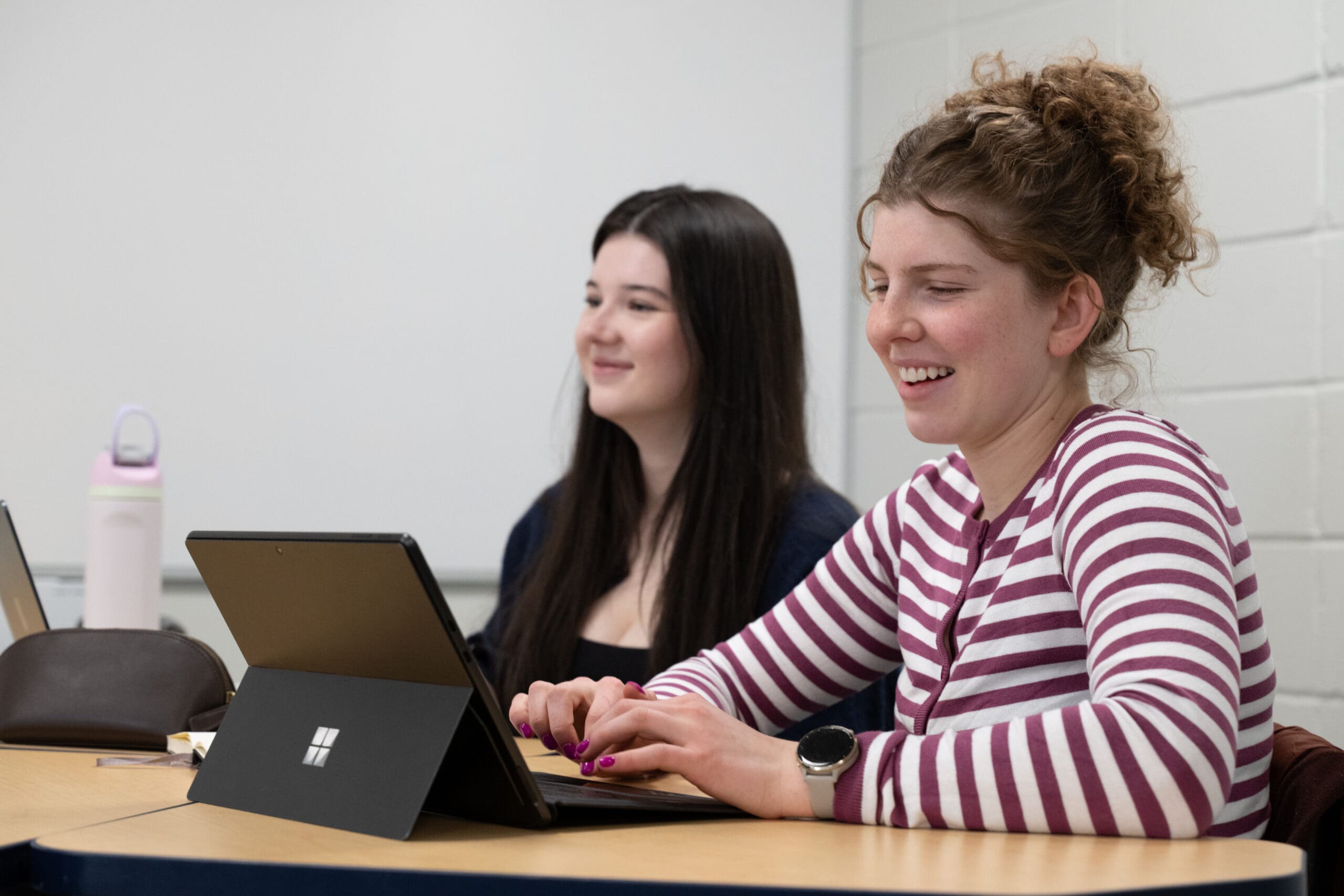 Deux jeunes femmes sont assises à une table, devant leurs ordinateurs portables. L'une porte un t-shirt rayé et sourit tout en tapant sur son clavier, tandis que l'autre, aux cheveux foncés, regarde vers l'avant de la salle. Toutes deux semblent concentrées sur leur travail.