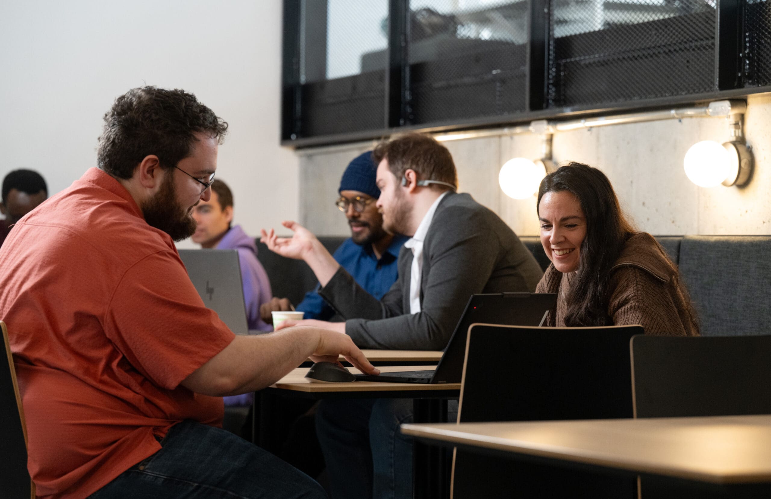 Un groupe de personnes est assis à des tables, un ordinateur portable devant elles, discutant et travaillant ensemble dans un espace de travail moderne. L'une d'entre elles, vêtue d'une chemise rouge, tape sur son clavier, tandis que les autres discutent et échangent des sourires.