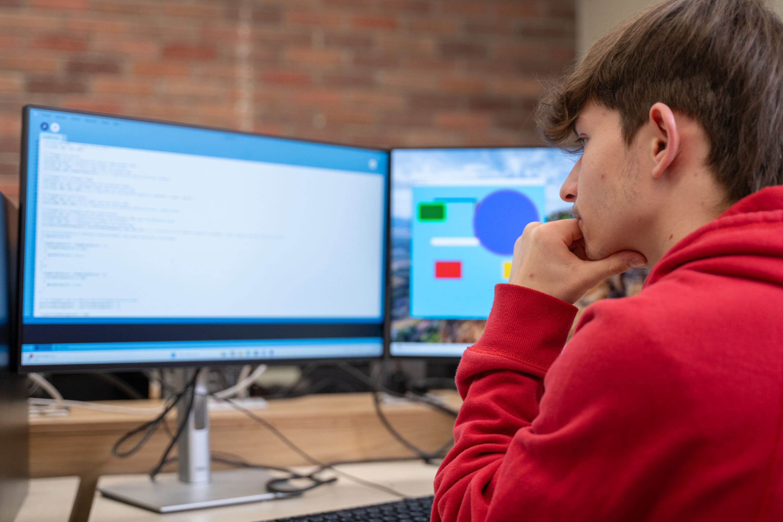 Une personne vêtue d'un sweat à capuche rouge est assise à un bureau ; elle observe d'un air pensif le code affiché sur l'écran d'un ordinateur, tandis qu'un autre écran, visible en arrière-plan, présente des graphiques colorés.