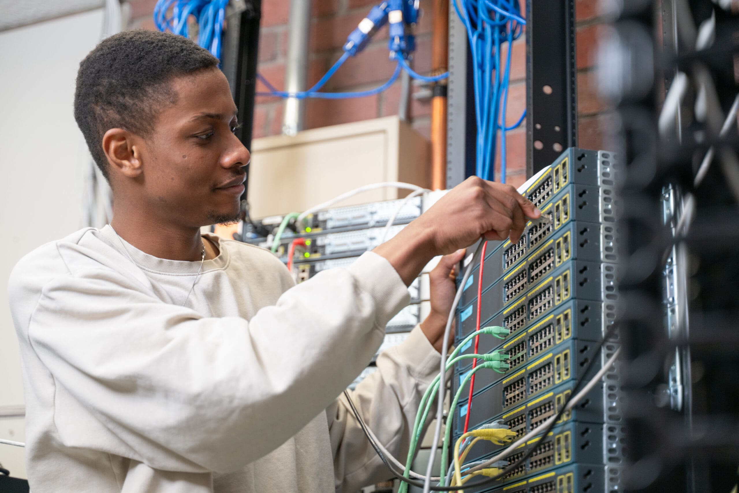Une personne raccorde des fils à des équipements réseau dans une salle de serveurs, au milieu d'une multitude de câbles et d'appareils électroniques.