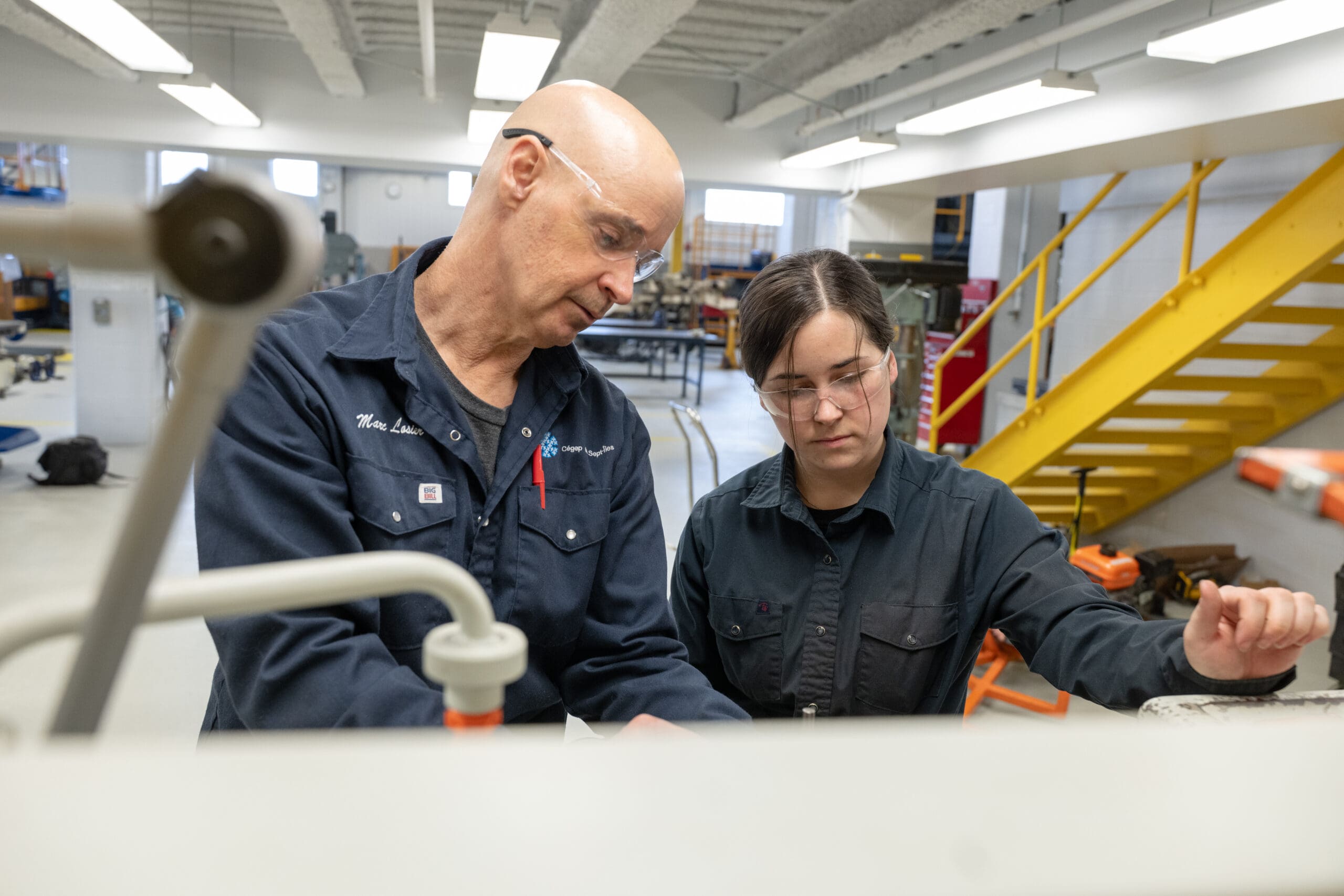 Deux personnes portant des lunettes de sécurité et des chemises de travail foncées examinent du matériel dans un atelier industriel. L'une d'elles désigne un élément de commande tandis que l'autre observe. On aperçoit, à l'arrière-plan, un escalier jaune et des machines.