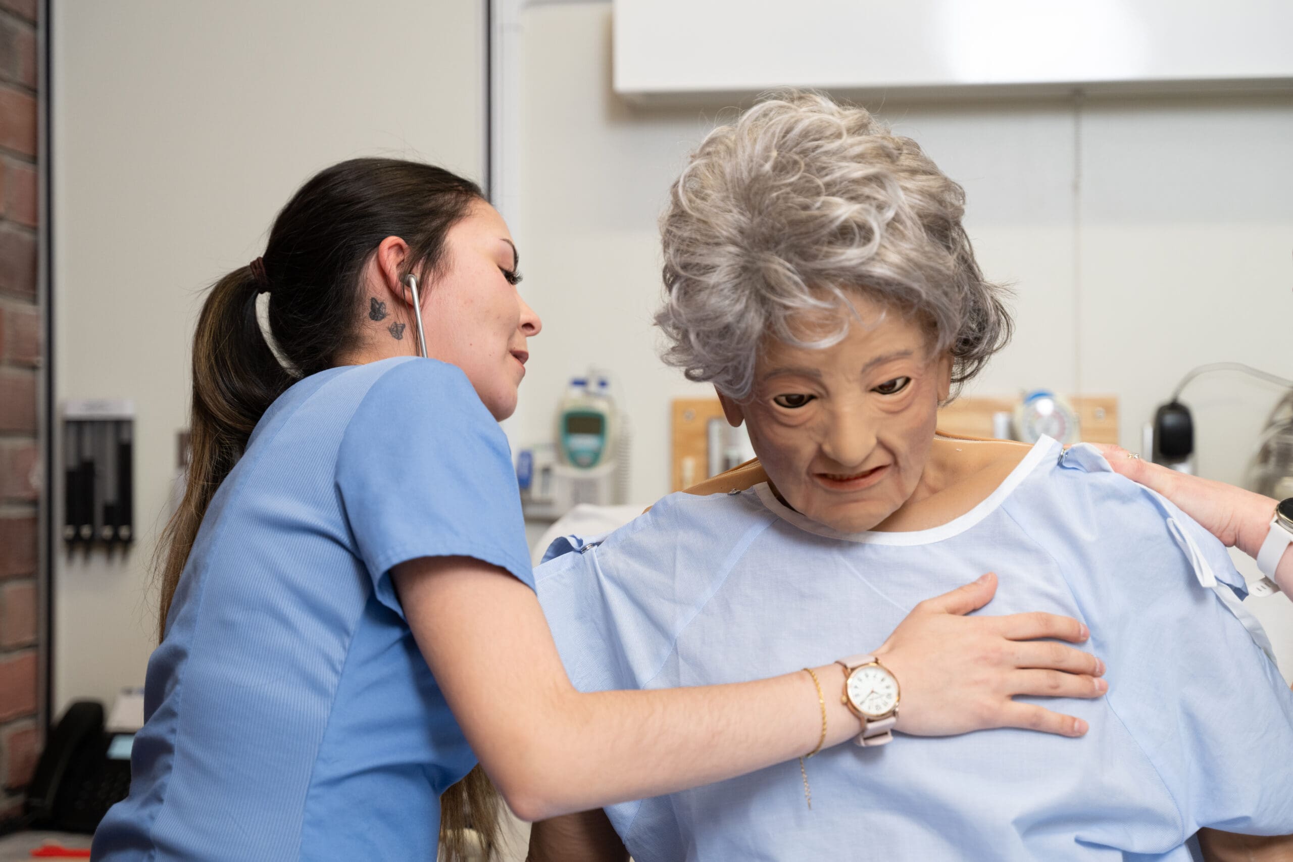 Dans le cadre d'une formation médicale, une infirmière vêtue d'une blouse bleue utilise un stéthoscope pour ausculter la poitrine d'un mannequin de patient vêtu d'une blouse d'hôpital.