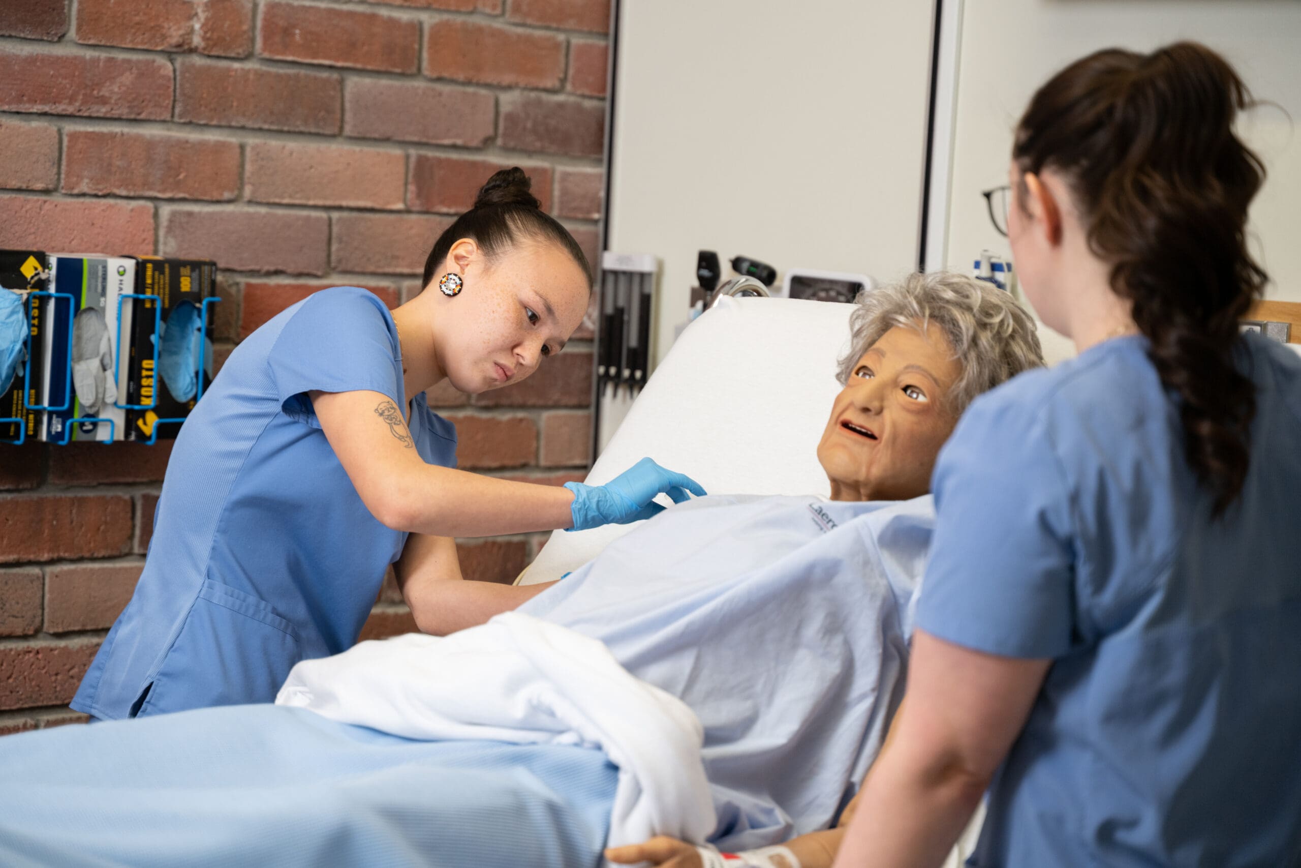 Deux personnes vêtues de blouses bleues s'occupent d'un mannequin médical allongé dans un lit d'hôpital, simulant ainsi la prise en charge d'un patient dans un cadre de formation clinique. Des instruments médicaux sont visibles sur le mur derrière elles.