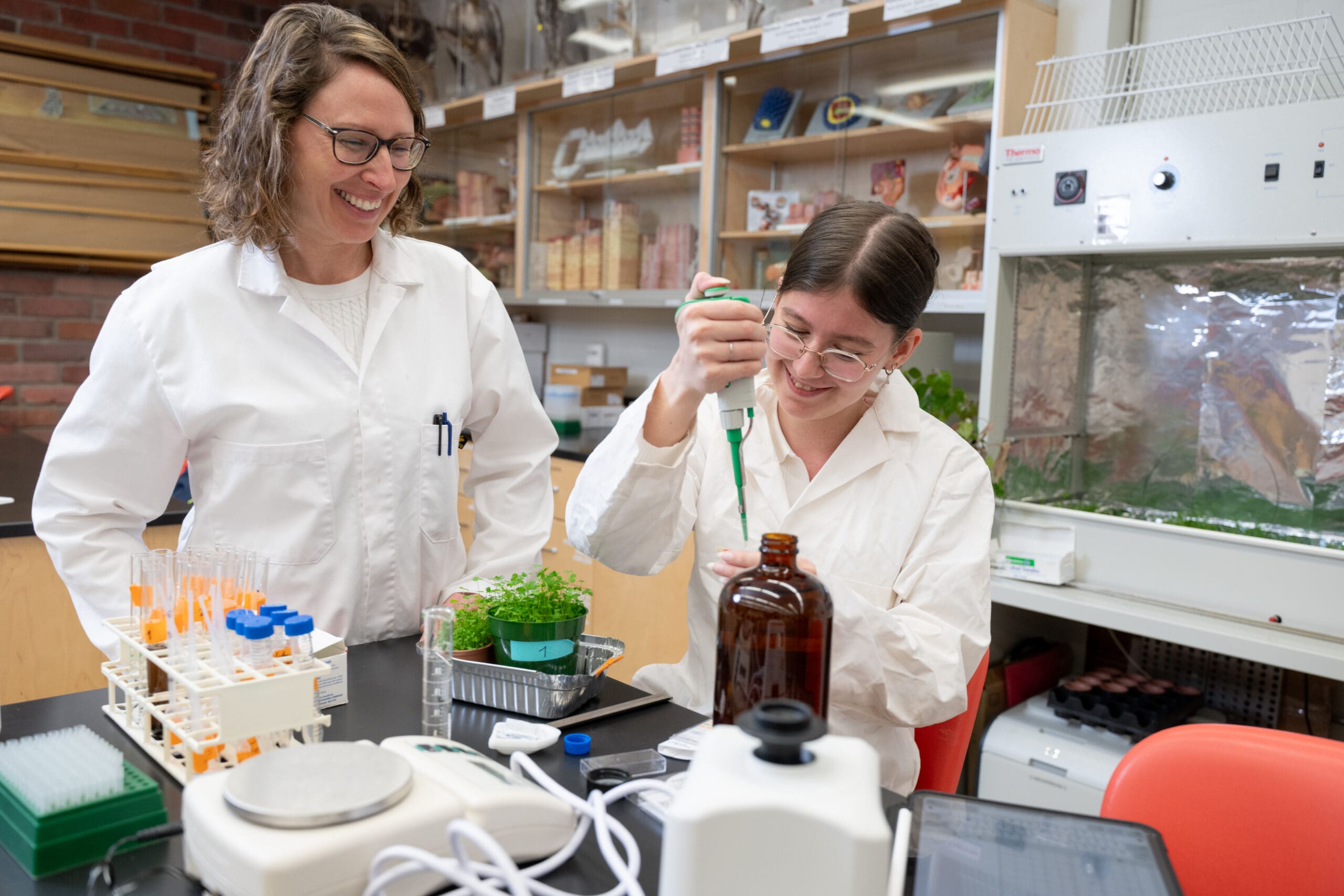 Deux scientifiques en blouse blanche travaillent dans un laboratoire. L'un d'eux sourit tandis que l'autre utilise une pipette. Des tubes à essai, des plantes et du matériel de laboratoire sont posés sur la table, et on aperçoit à l'arrière-plan des étagères remplies de fournitures.