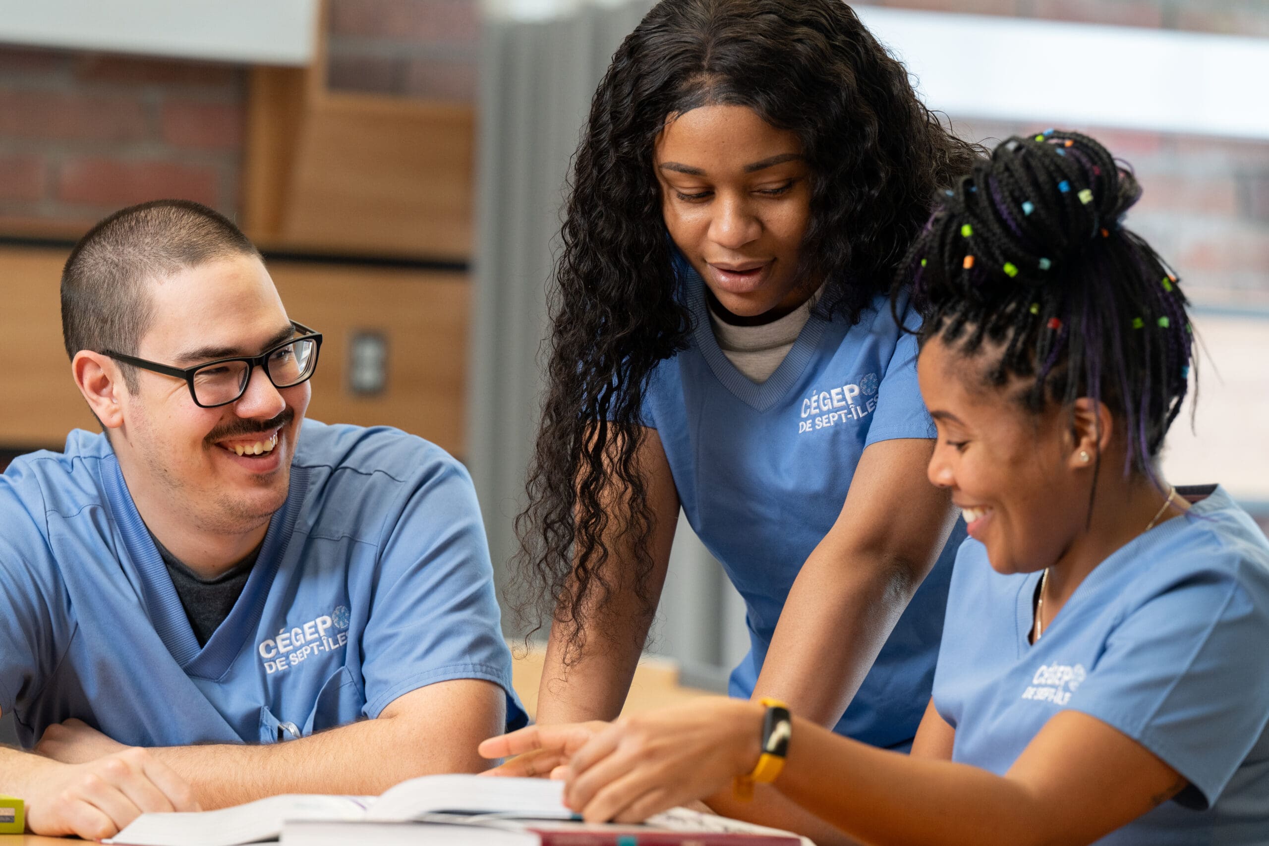 Trois étudiants vêtus de blouses bleues étudient ensemble autour d'une table ; ils sourient et discutent d'un sujet en consultant un livre ouvert. Ils semblent concentrés et travaillent en collaboration dans un intérieur lumineux.