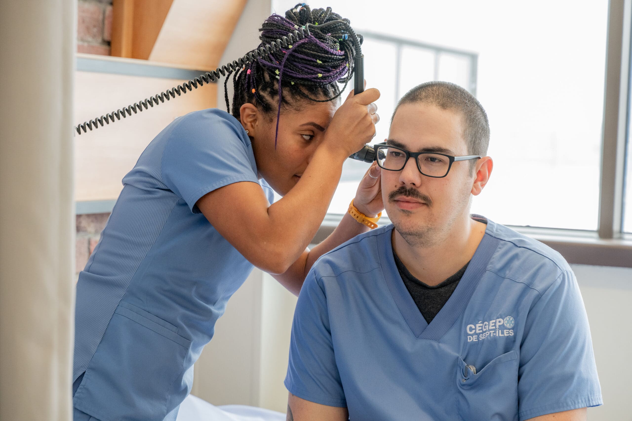 Un professionnel de santé vêtu d'une blouse bleue examine l'oreille d'un collègue à l'aide d'un otoscope. Tous deux semblent concentrés, et la pièce est baignée de lumière naturelle.