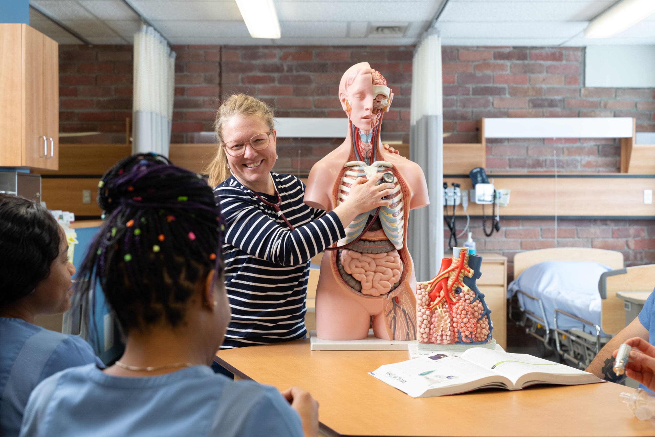 Une femme dispense un cours à des élèves à l'aide d'un modèle anatomique humain dans une salle de classe. Deux élèves l'écoutent, tandis qu'un manuel d'anatomie et un modèle de cœur sont posés sur la table.