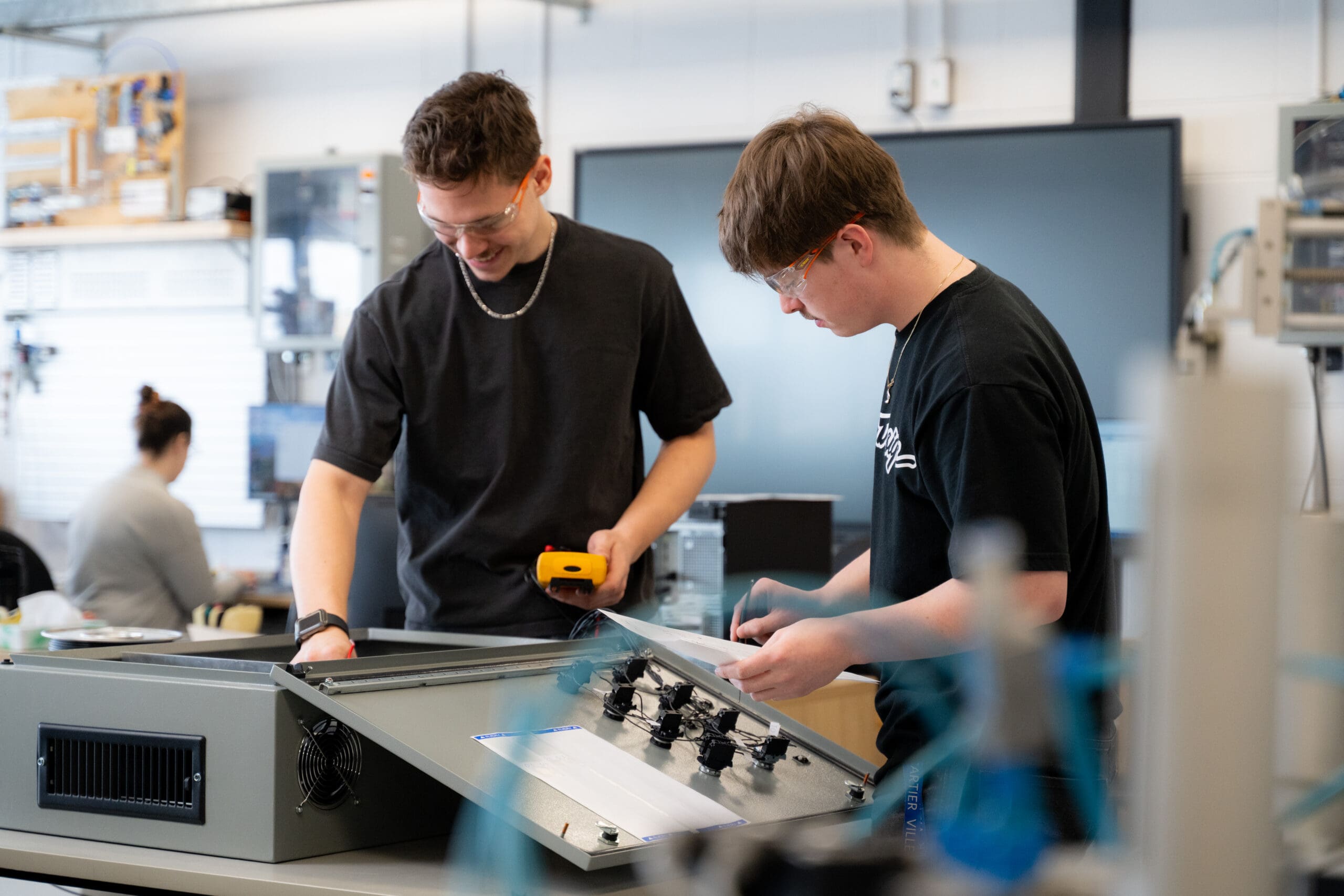 Deux personnes portant des lunettes de sécurité travaillent sur un tableau de commande électronique ouvert dans un laboratoire ; elles utilisent des outils et examinent des composants.