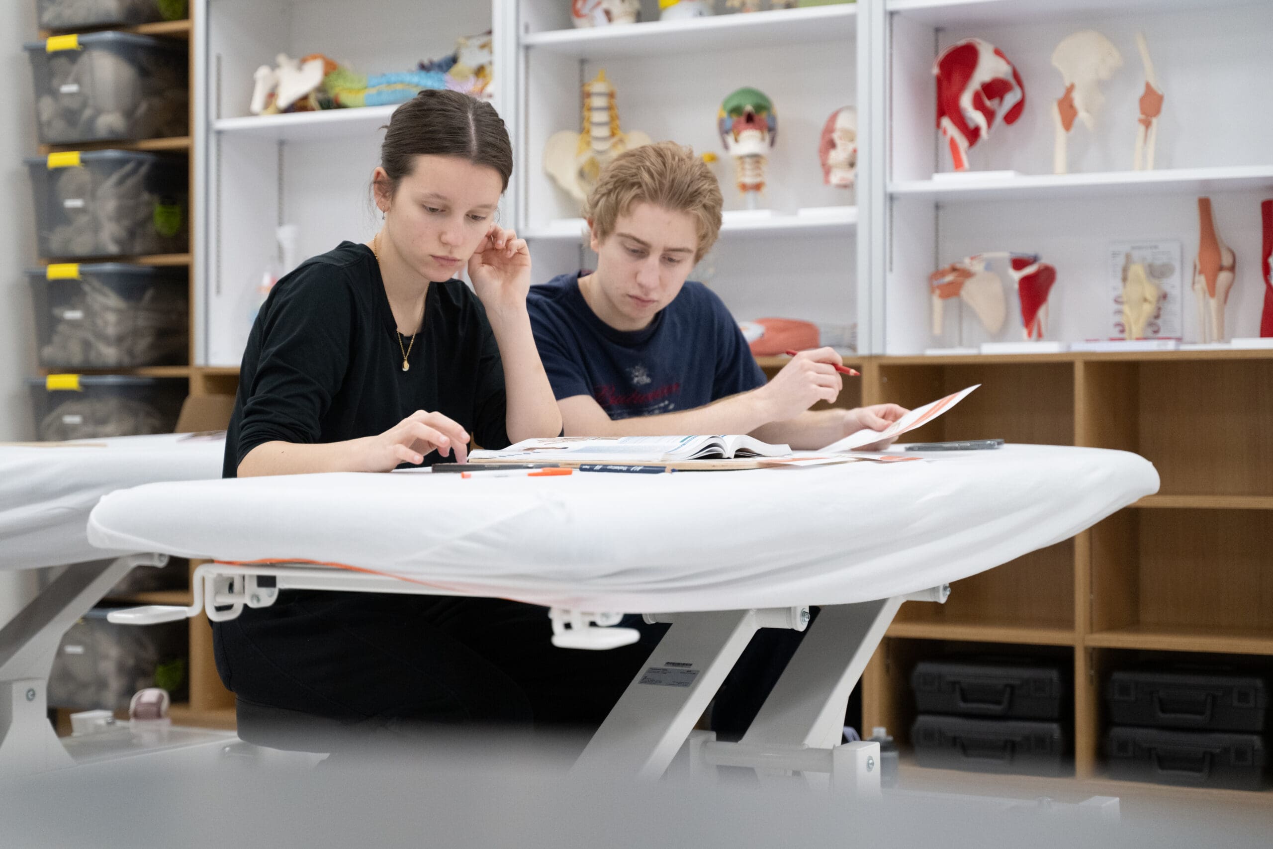 Deux élèves sont assis à une table et étudient ensemble des manuels d'anatomie dans une salle de classe où se trouvent, sur les étagères derrière eux, des modèles anatomiques et des squelettes.