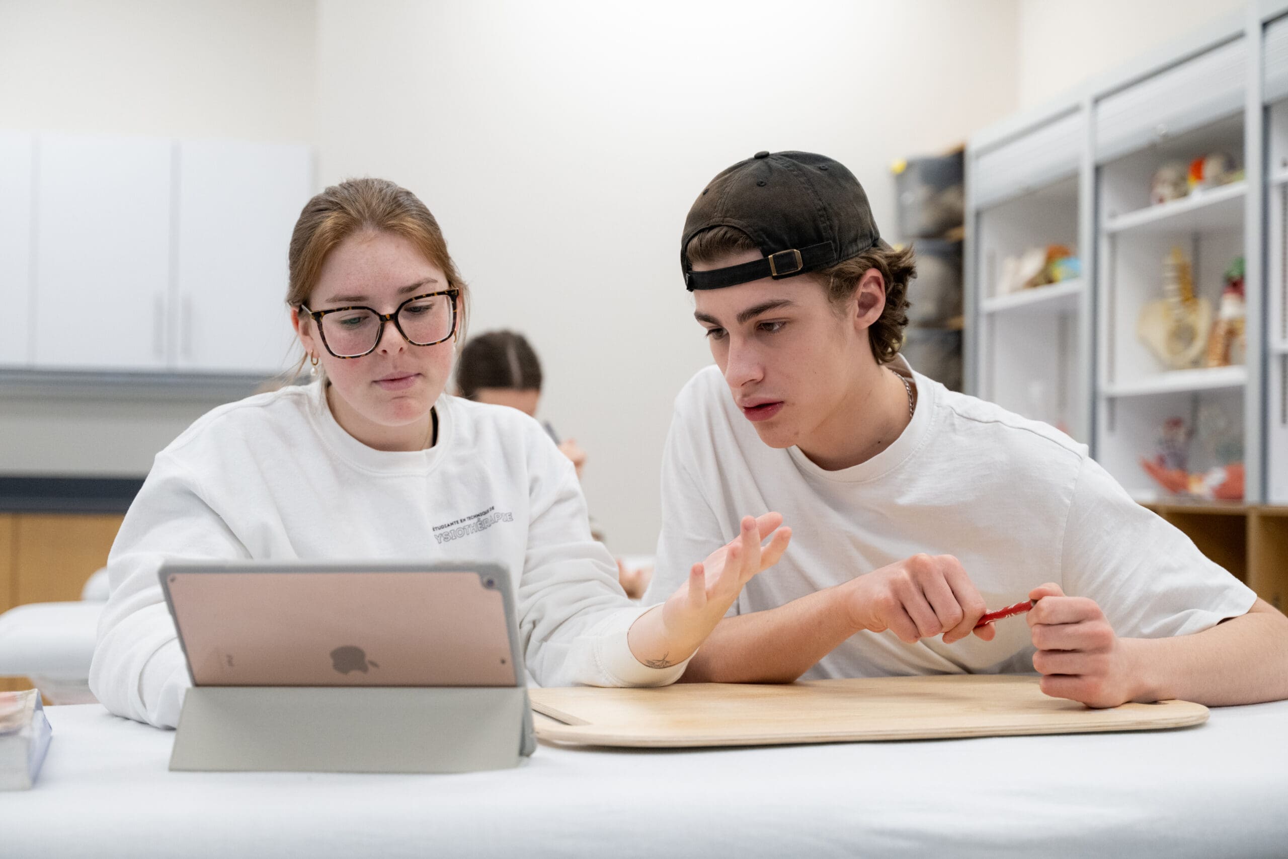 Deux étudiants sont assis à une table et regardent ensemble une tablette. L'un d'eux fait des gestes de la main tandis que l'autre tient un stylet. On aperçoit à l'arrière-plan des étagères sur lesquelles sont disposés des modèles anatomiques.