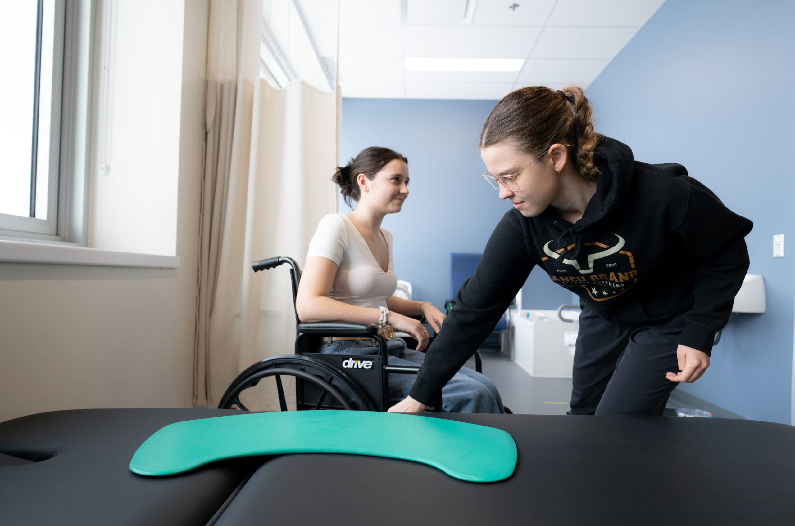 Dans un établissement médical ou de rééducation, une femme en fauteuil roulant observe une autre femme en train de placer une planche de transfert verte sur une table recouverte d’un tapis noir.