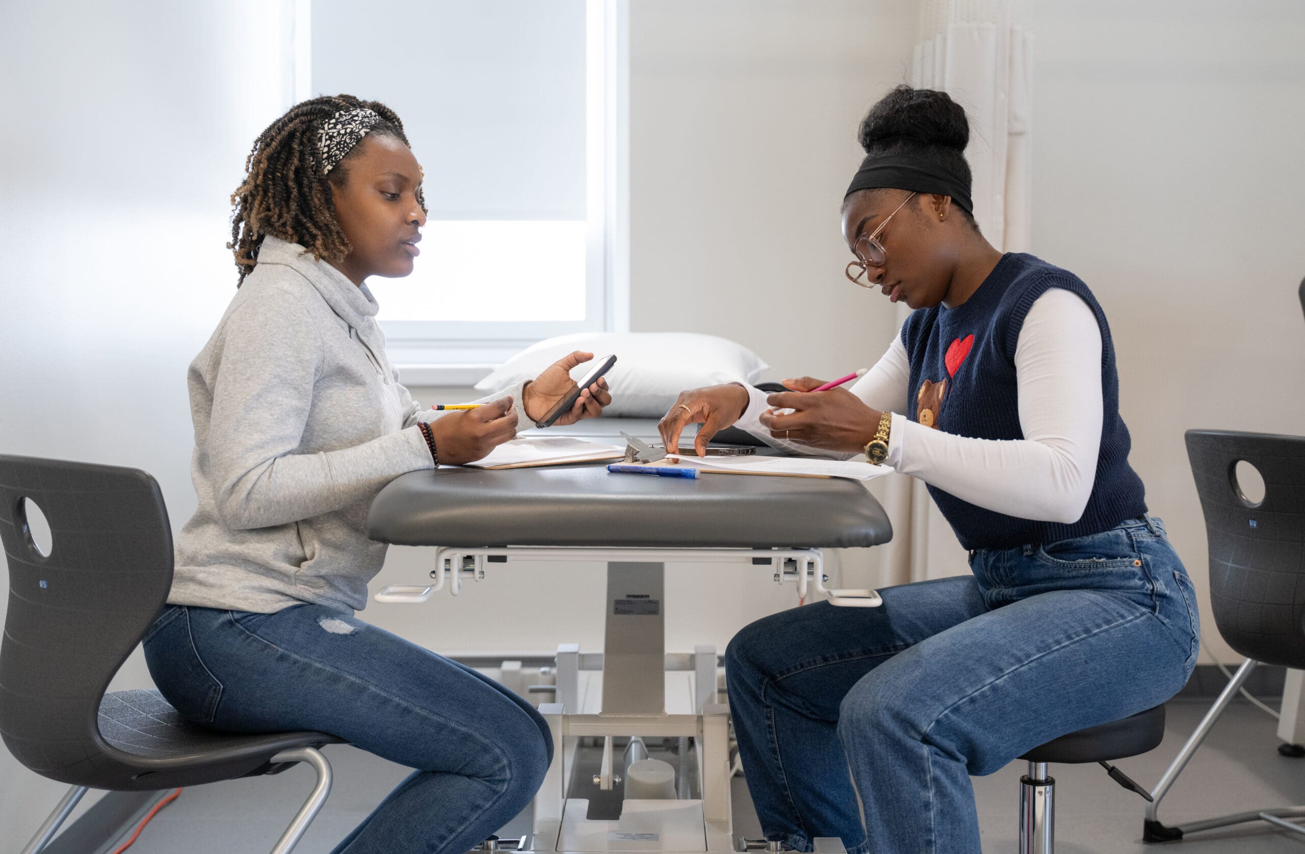 Deux femmes sont assises face à face à une table, les yeux rivés sur leurs téléphones et prenant des notes, avec des carnets et des feuilles étalés devant elles dans une pièce lumineuse.