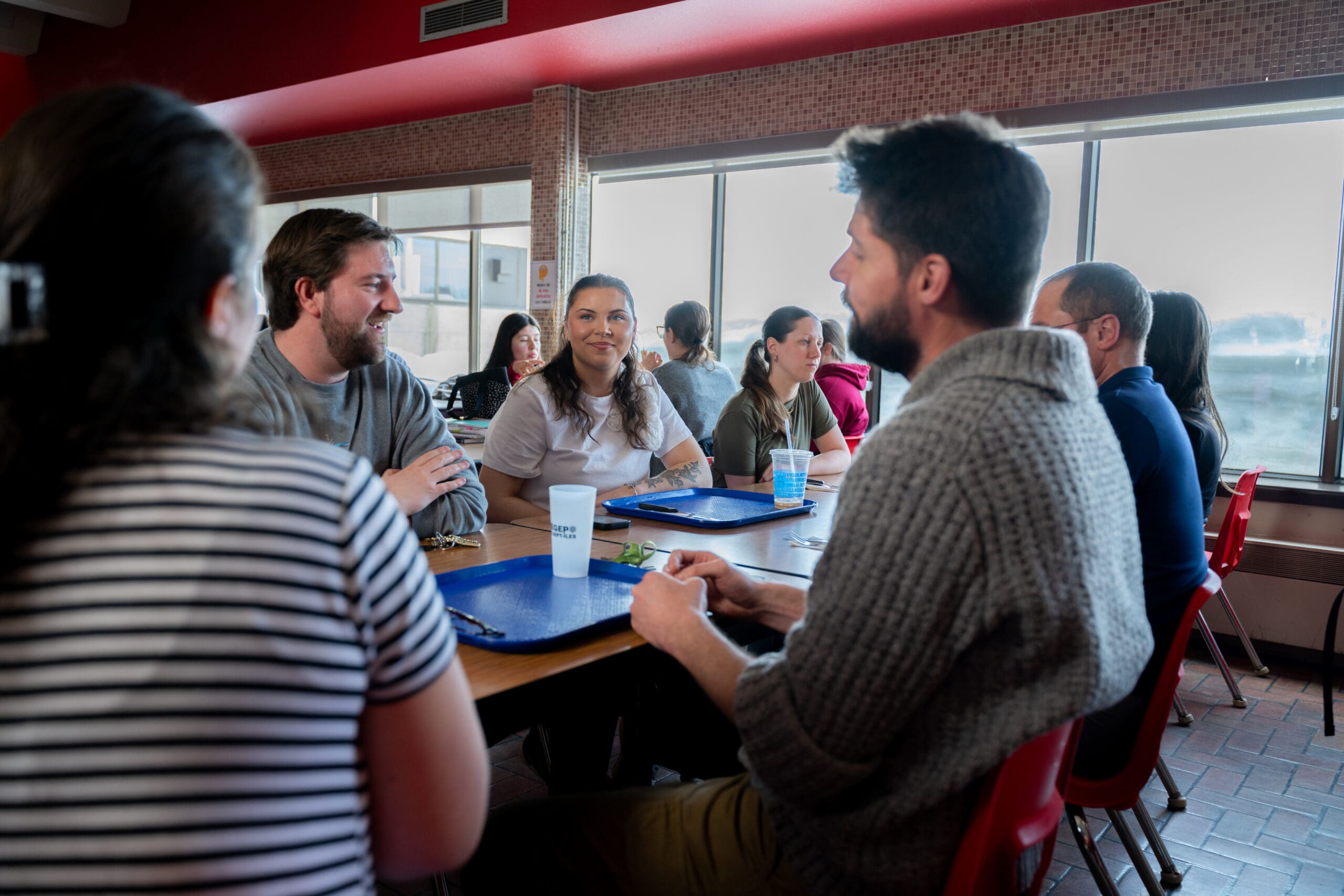 Un groupe de personnes est assis à une table de la cafétéria, des plateaux bleus à la main ; elles mangent et discutent, tandis que la lumière du soleil pénètre à flots par les grandes fenêtres situées derrière elles.
