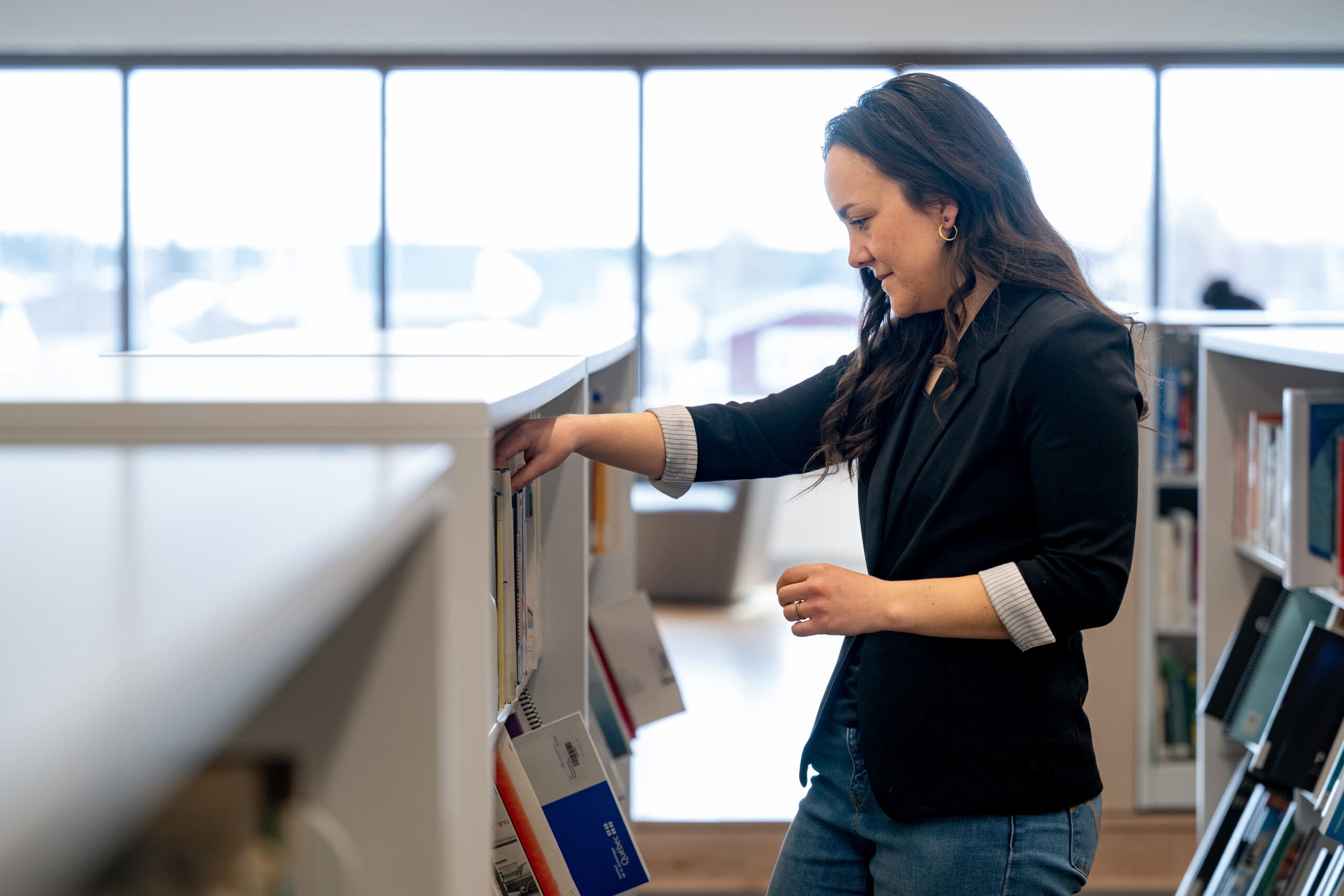 Une femme vêtue d'un blazer noir et d'un jean se tient debout dans une bibliothèque, en train de prendre un livre sur une étagère, avec en arrière-plan de grandes fenêtres laissant entrer la lumière naturelle.