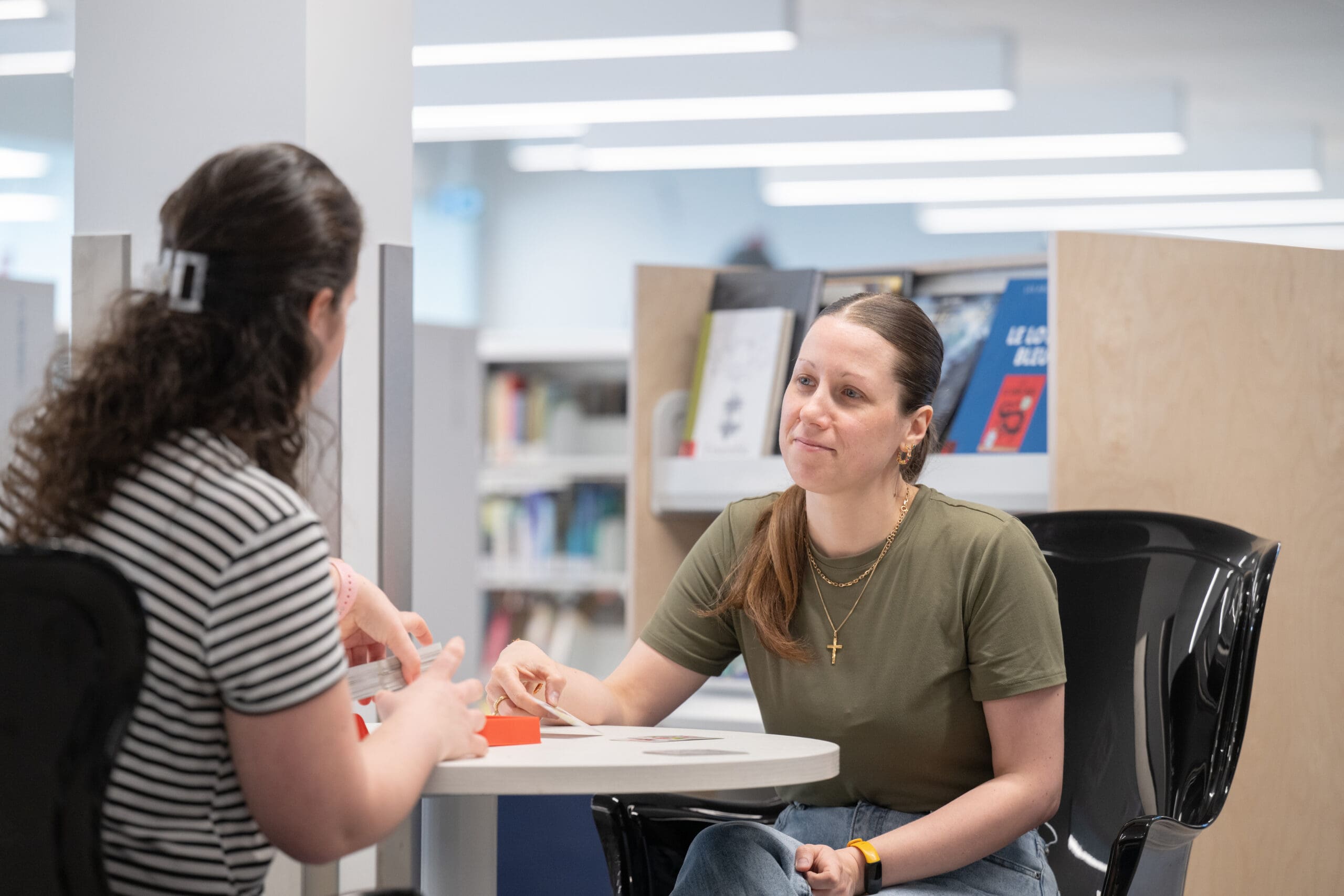 Deux femmes sont assises face à face à une table ronde dans une bibliothèque, en pleine conversation. L'une porte un t-shirt vert et a les cheveux attachés en arrière ; l'autre porte un t-shirt rayé. On aperçoit des étagères à l'arrière-plan.