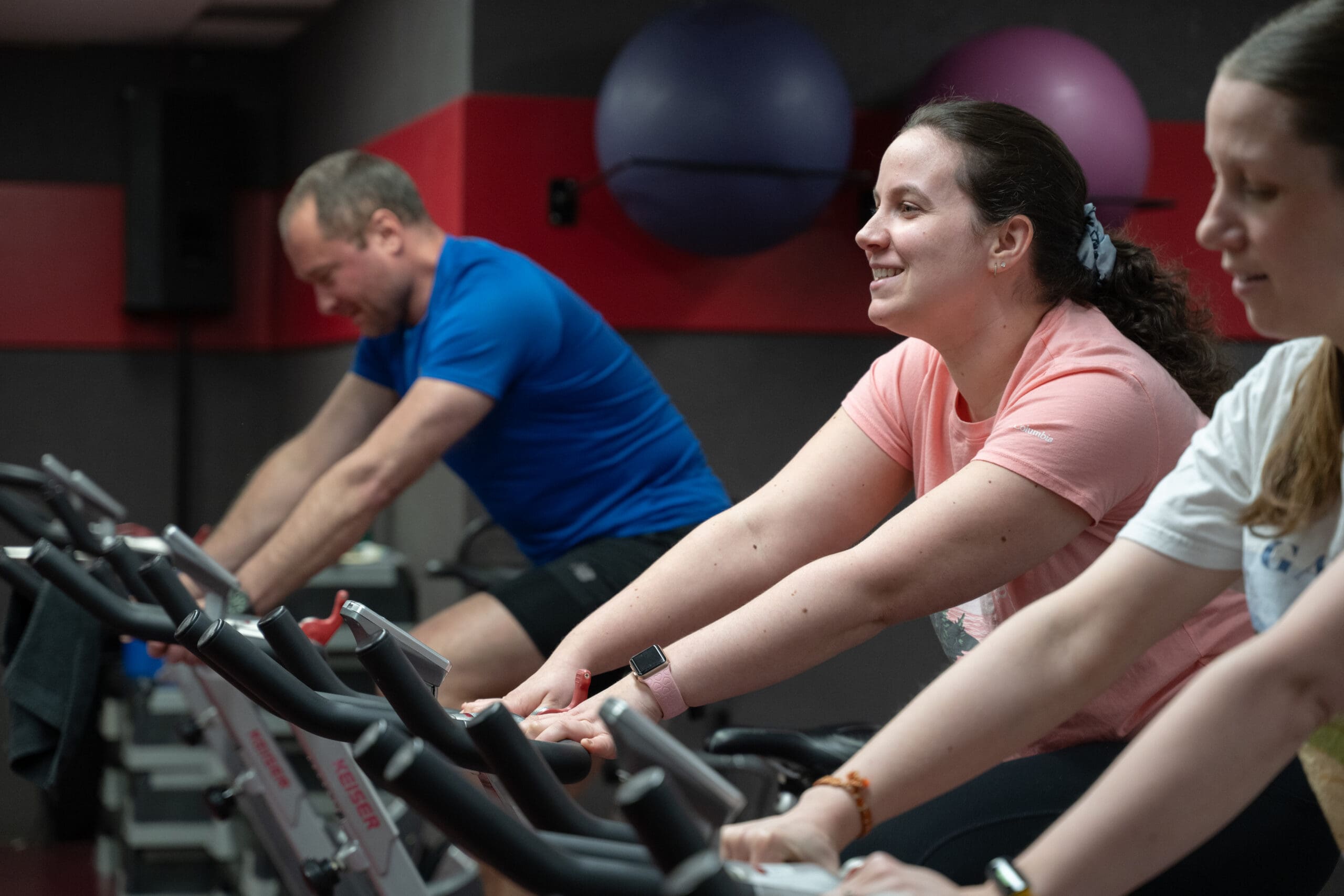 Trois personnes font du vélo d'appartement dans une salle de sport ; elles sont concentrées et souriantes pendant leur entraînement. Deux gros ballons d'exercice sont accrochés au mur, à l'arrière-plan.