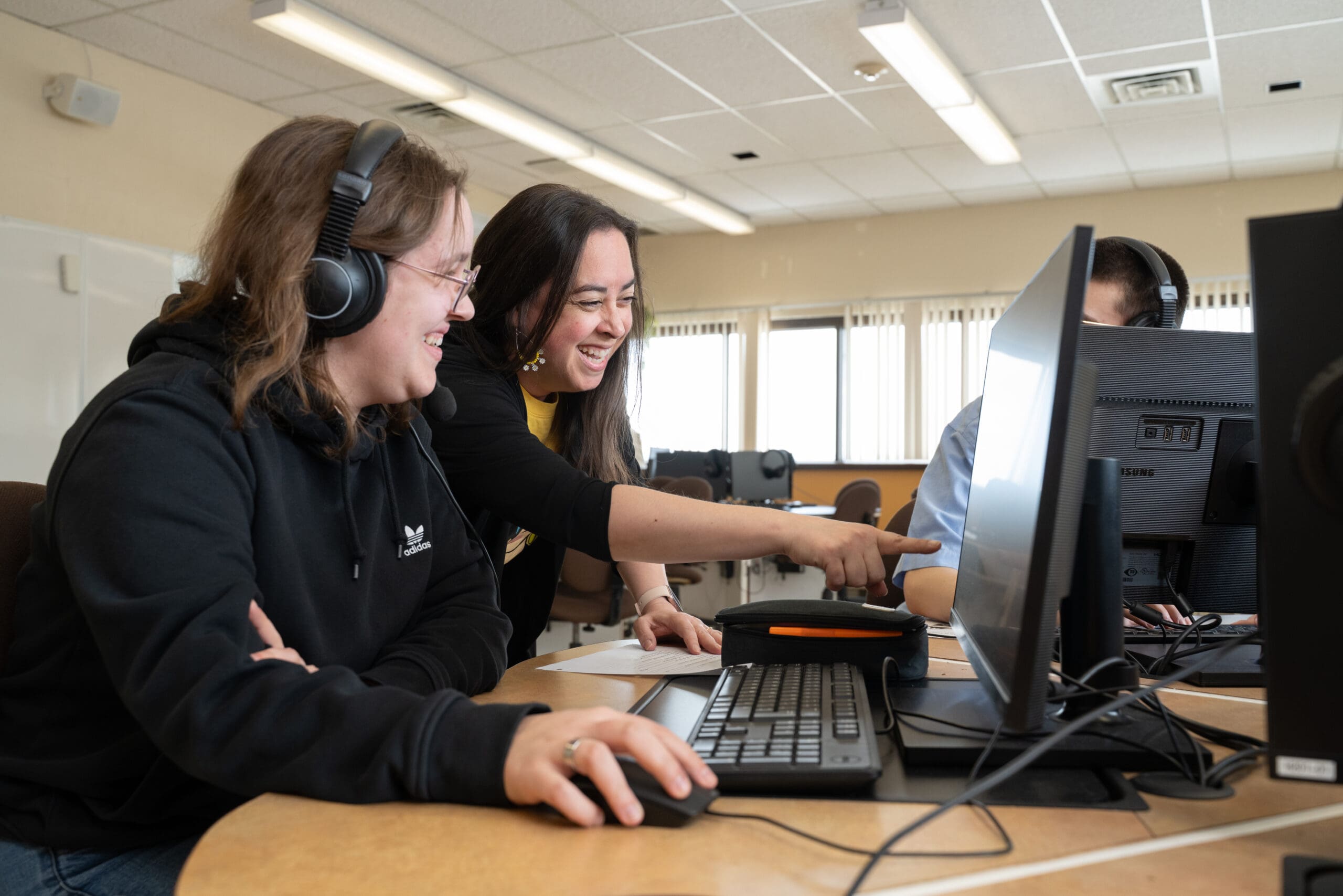Deux personnes souriantes regardant l'écran d'un ordinateur, dont l'une pointe du doigt le moniteur, ce qui suggère une situation de collaboration ou d'enseignement dans une salle de classe ou un bureau.