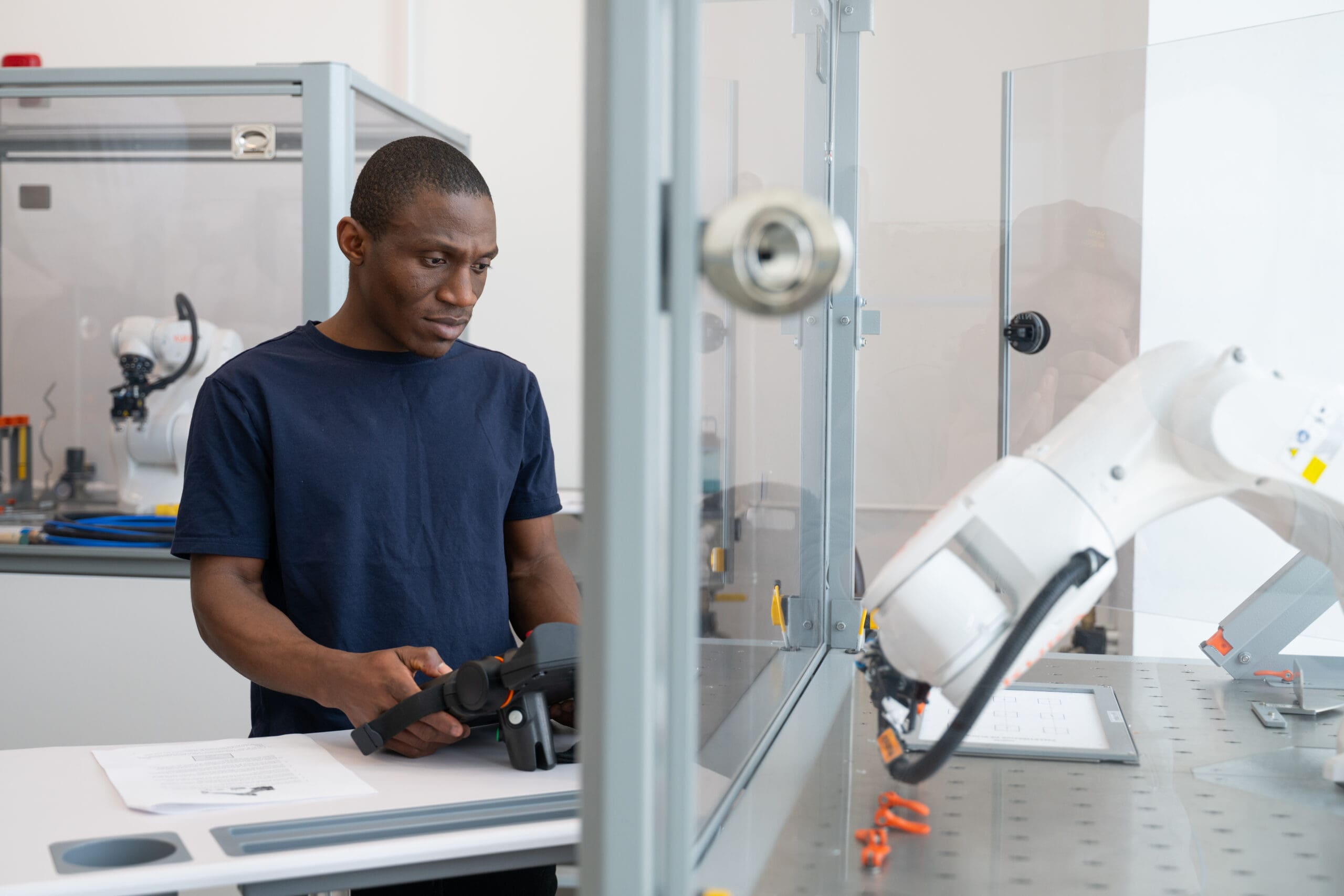 Dans un laboratoire moderne, où l'on aperçoit divers équipements tout autour de lui, un homme vêtu d'une chemise bleu marine manœuvre un bras robotisé à l'aide d'un dispositif de commande.
