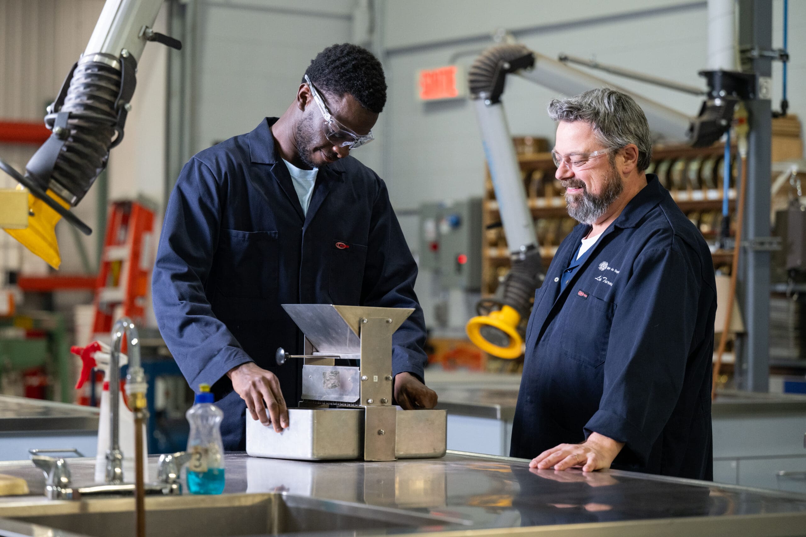 Deux hommes en blouse de laboratoire travaillent ensemble dans un atelier, examinant un appareil métallique posé sur une table. L'un d'eux règle l'appareil tandis que l'autre l'observe. On aperçoit du matériel industriel et des étagères à l'arrière-plan.