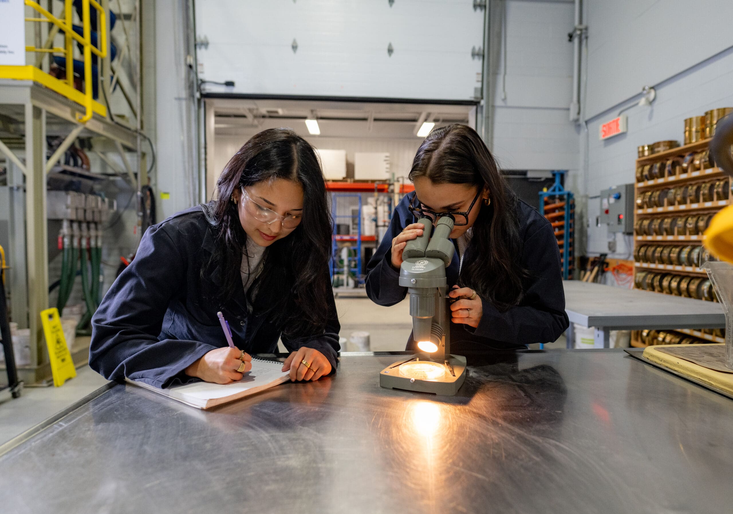 Deux femmes en blouse de laboratoire travaillent dans un laboratoire ; l'une prend des notes sur un bloc-notes, tandis que l'autre observe au microscope posé sur une table métallique.