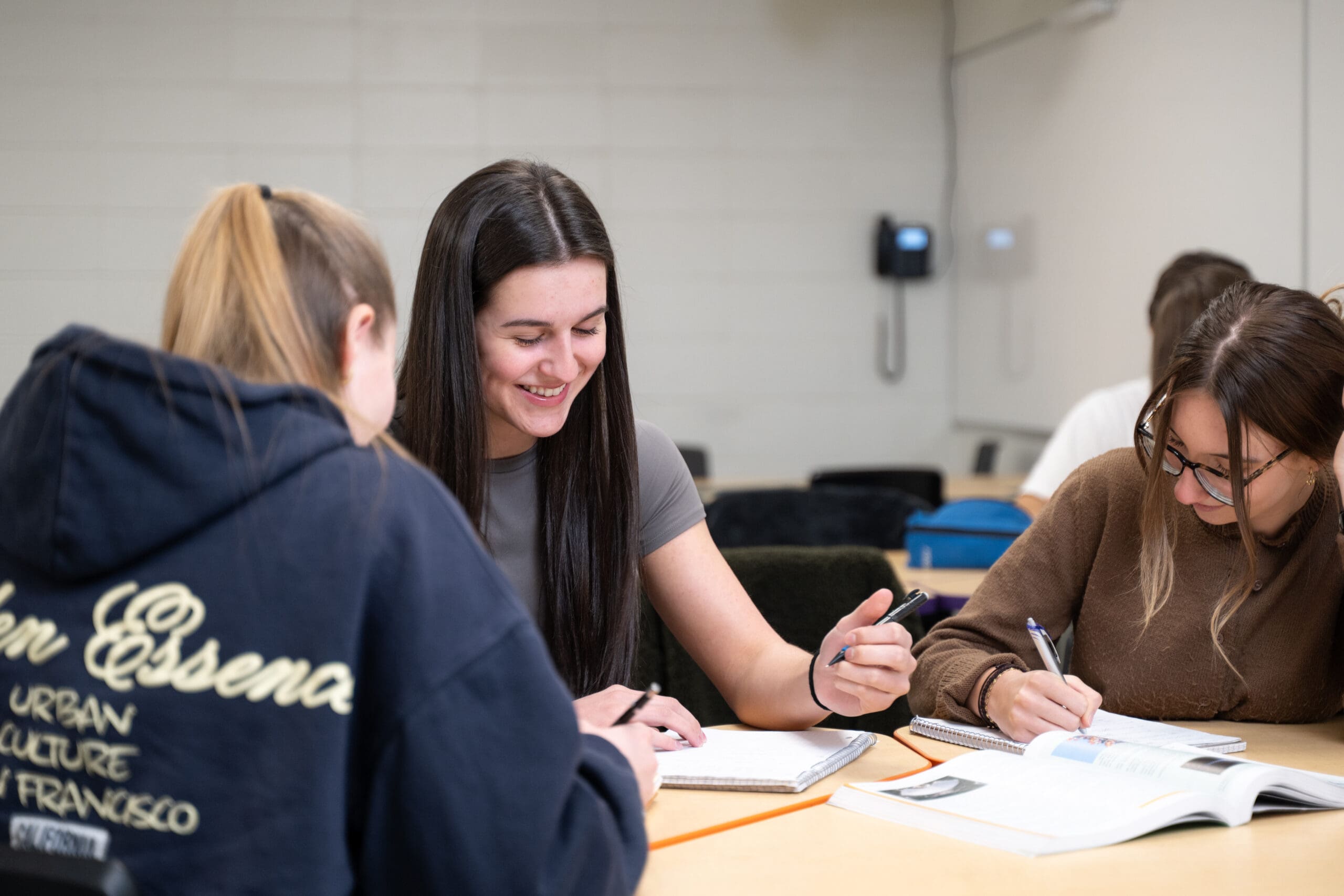 Trois jeunes femmes sont assises à une table, souriantes, en train d'écrire dans leurs cahiers, entourées de manuels scolaires, dans une salle de classe.