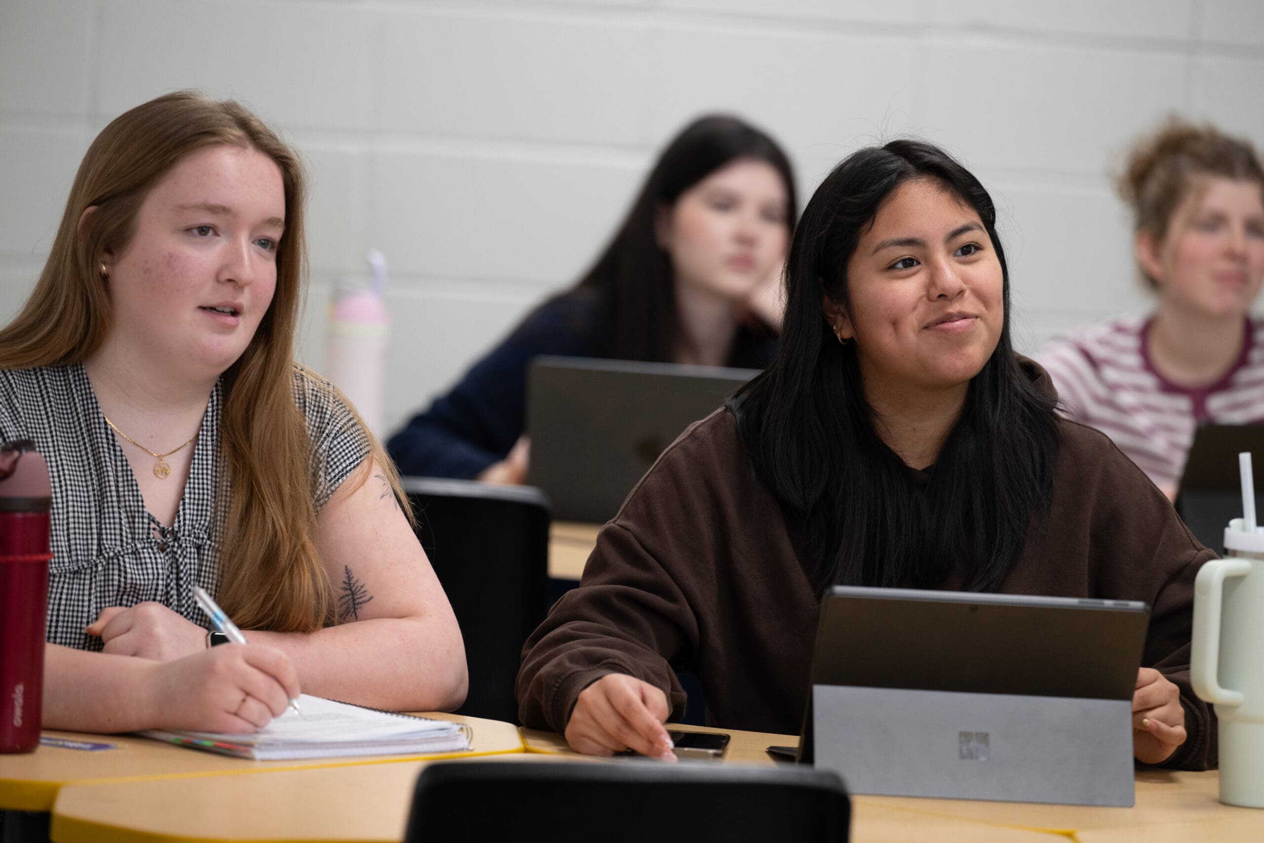Quatre élèves sont assis à leurs pupitres dans une salle de classe, équipés de tablettes et d'ordinateurs portables. Les deux élèves assis au premier rang participent activement au cours et prennent des notes, tandis que les deux autres, assis à l'arrière, écoutent attentivement.