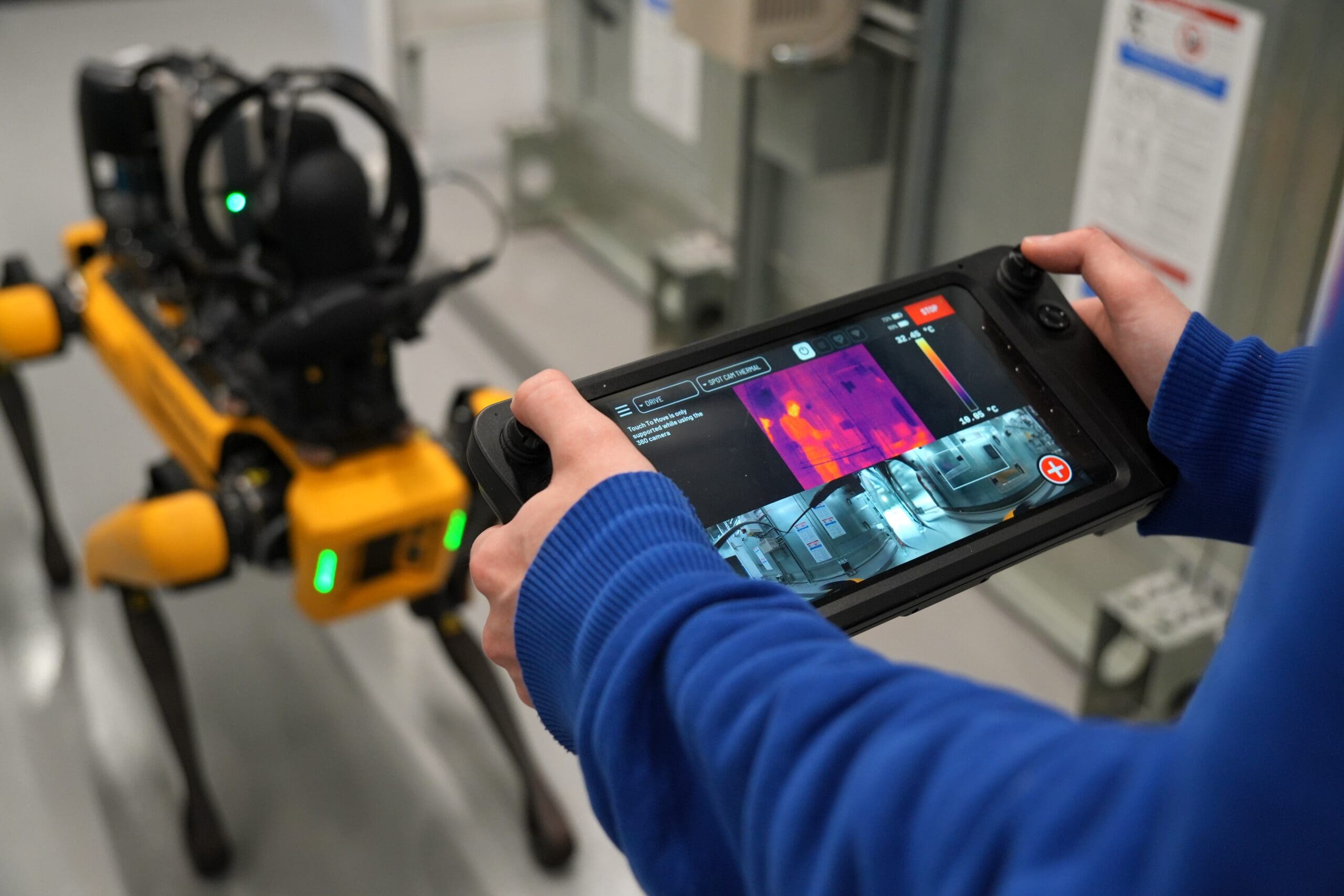 Une personne pilote un chien robot jaune à l'aide d'une tablette, qui affiche les images captées par la caméra thermique et la caméra classique du robot, du point de vue de ce dernier, dans un environnement intérieur.
