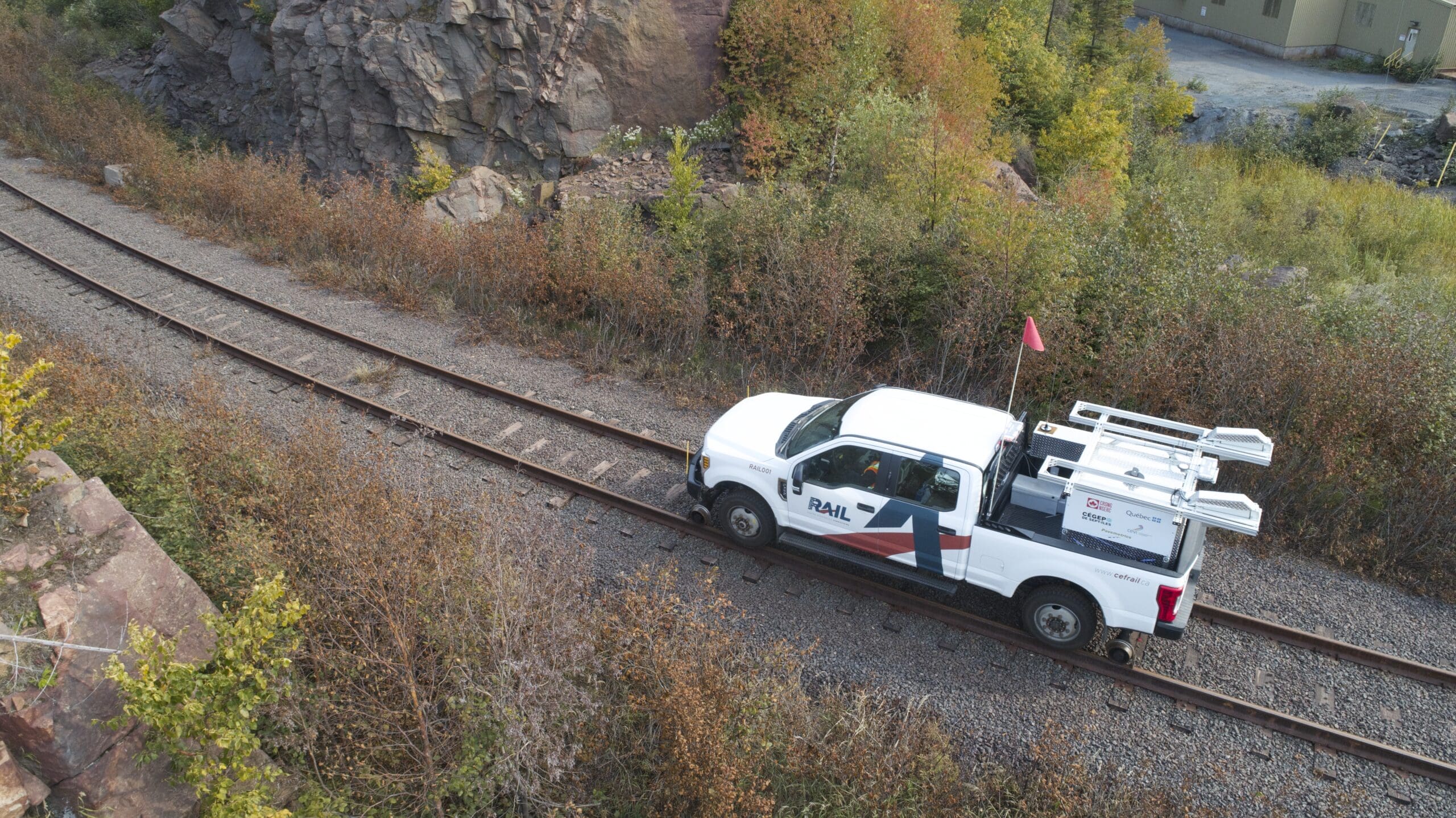 Une camionnette blanche de service, chargée de matériel à l'arrière, roule le long d'une voie ferrée, au milieu des rochers et d'une végétation verdoyante et dense.
