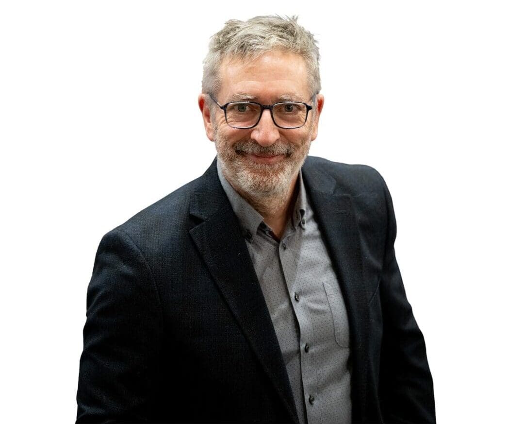 Un homme d'âge mûr, aux cheveux gris, portant des lunettes et une barbe, vêtu d'une veste de costume sombre et d'une chemise à motifs gris clair, sourit en regardant l'appareil photo devant un fond blanc uni.