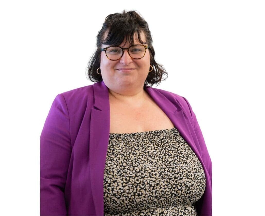 Une personne aux cheveux foncés et portant des lunettes, vêtue d'un blazer violet et d'un haut à motifs noir et blanc, sourit à la caméra devant un fond blanc uni.
