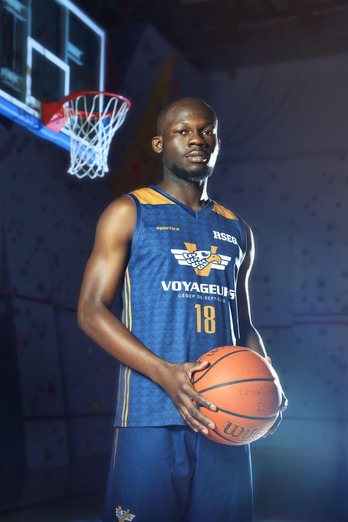 Un basketteur, vêtu d'un maillot bleu « Voyageurs », se tient à l'intérieur, un ballon de basket à la main. On aperçoit un panier de basket à l'arrière-plan, sous un éclairage spectaculaire.