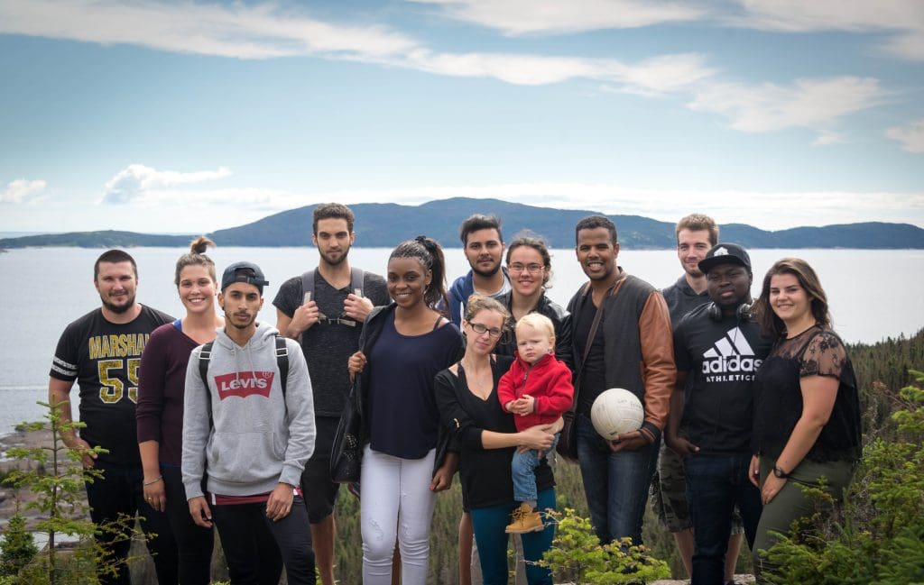 Un groupe hétéroclite de douze personnes, dont un enfant, pose en plein air sur une colline, avec de la verdure, de l'eau et des montagnes en arrière-plan. Un homme tient un ballon de football. Le ciel est partiellement nuageux.