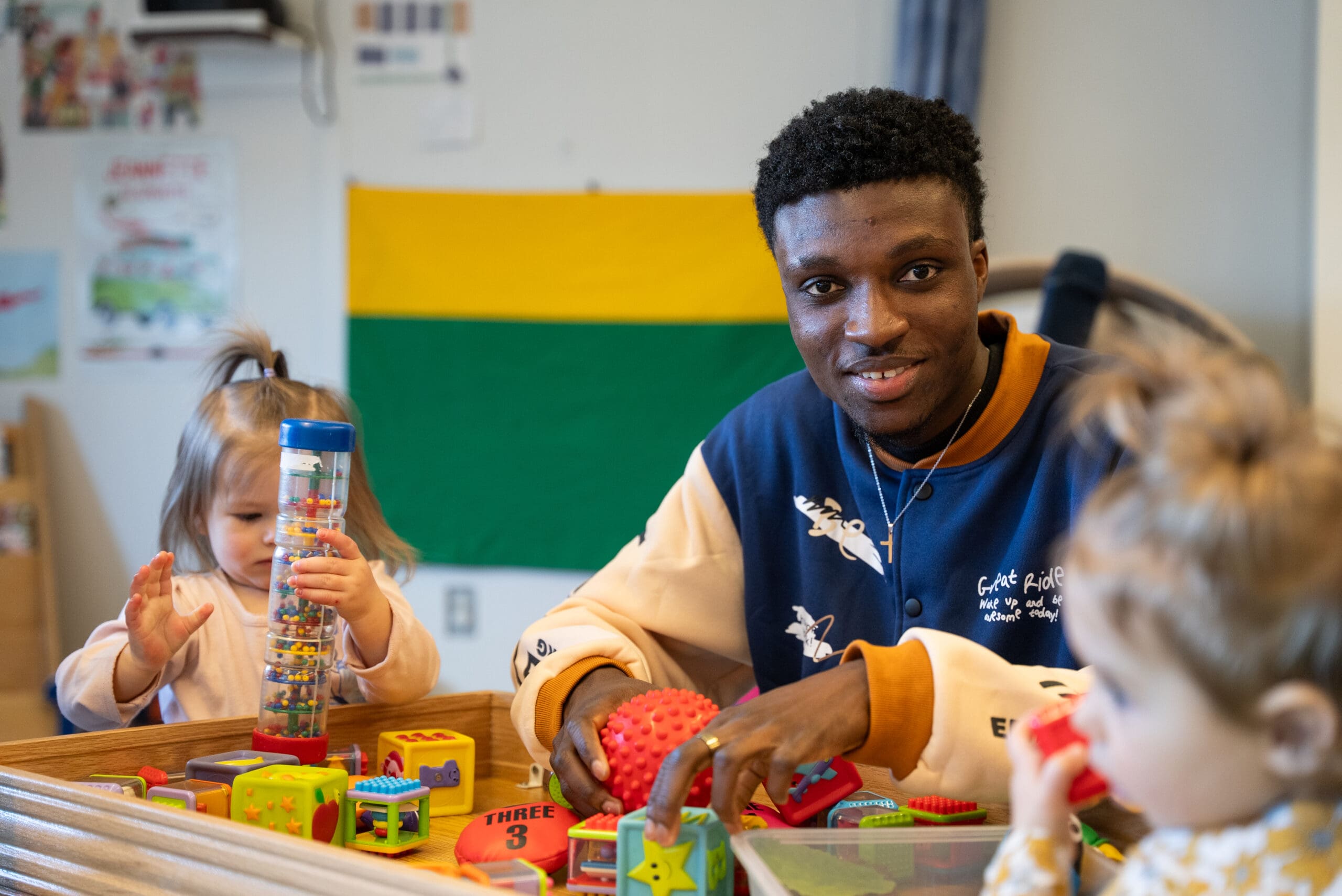 Un homme sourit tout en jouant avec deux jeunes enfants autour d'une table couverte de jouets et de blocs colorés. Un drapeau vert, jaune et noir est accroché au mur derrière eux. Les enfants sont absorbés par leurs jouets.
