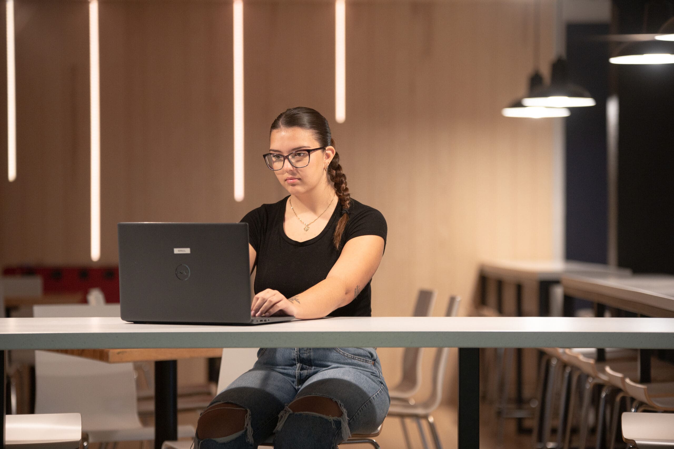 Une femme portant des lunettes et une tresse est assise à une longue table, devant un ordinateur portable, dans un espace moderne et bien éclairé, aux murs en bois et orné de suspensions lumineuses.