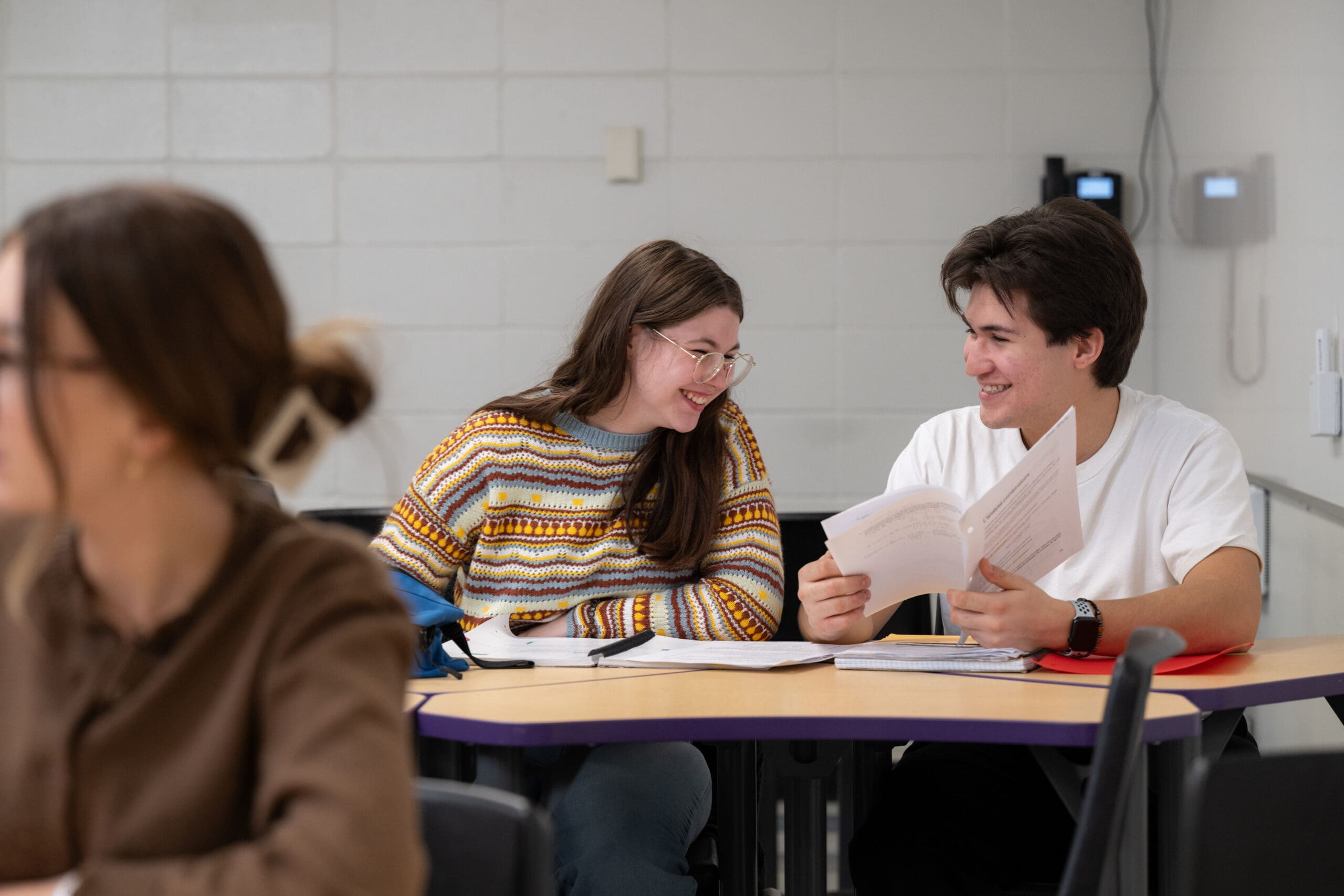 Deux élèves assis à une table dans une salle de classe sourient et se regardent tout en tenant des feuilles. Un autre élève apparaît flou au premier plan.