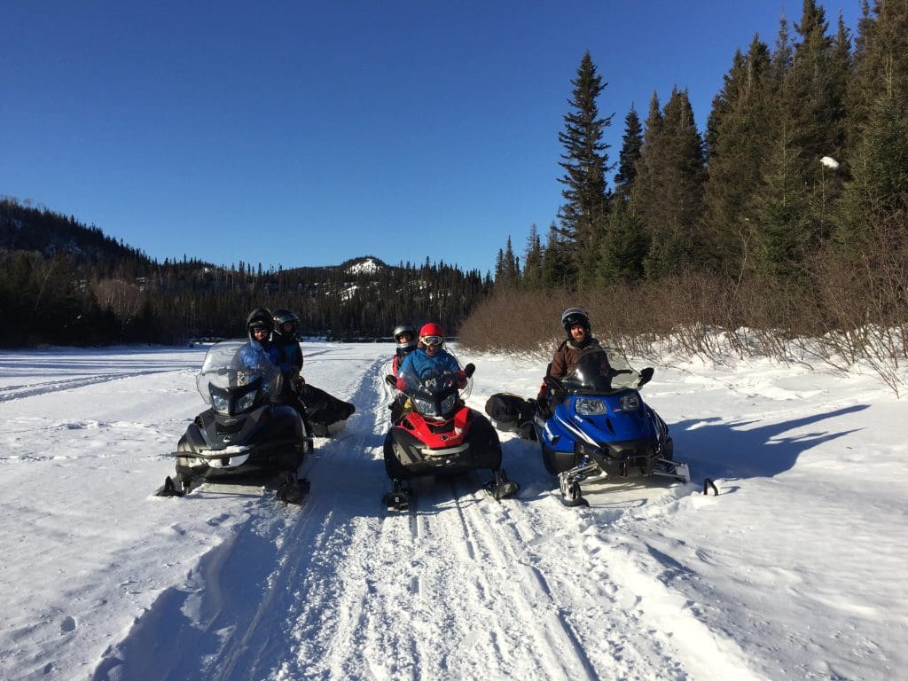 Quatre personnes vêtues de tenues d'hiver sont assises sur des motoneiges garées sur une piste enneigée, entourées d'un paysage recouvert de neige, d'arbres à feuilles persistantes et de collines lointaines, sous un ciel bleu limpide.