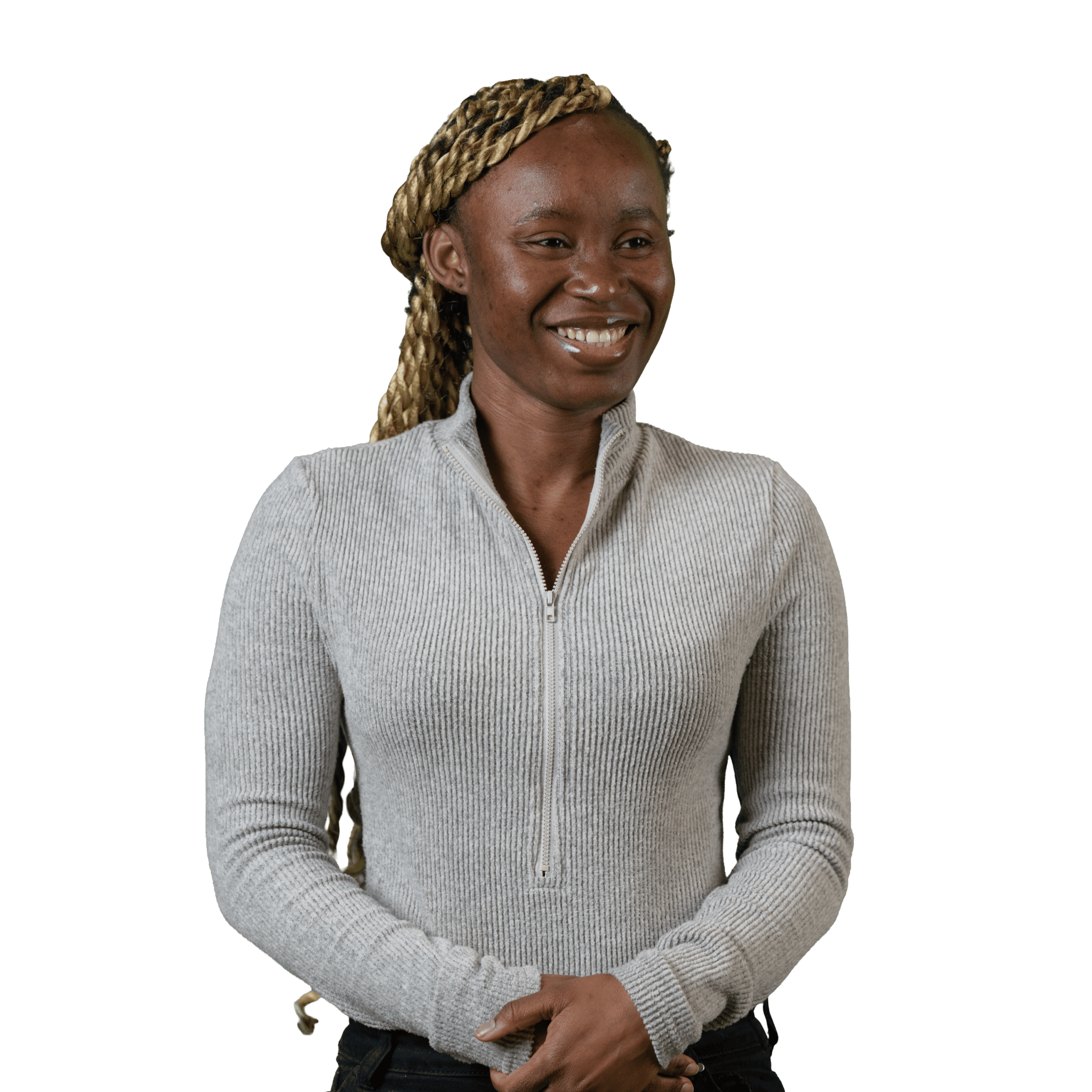 Une femme aux cheveux tressés, vêtue d'un pull à col zippé gris clair à côtes, se tient debout devant un fond blanc, souriante, le regard légèrement tourné sur le côté, les mains jointes devant elle.