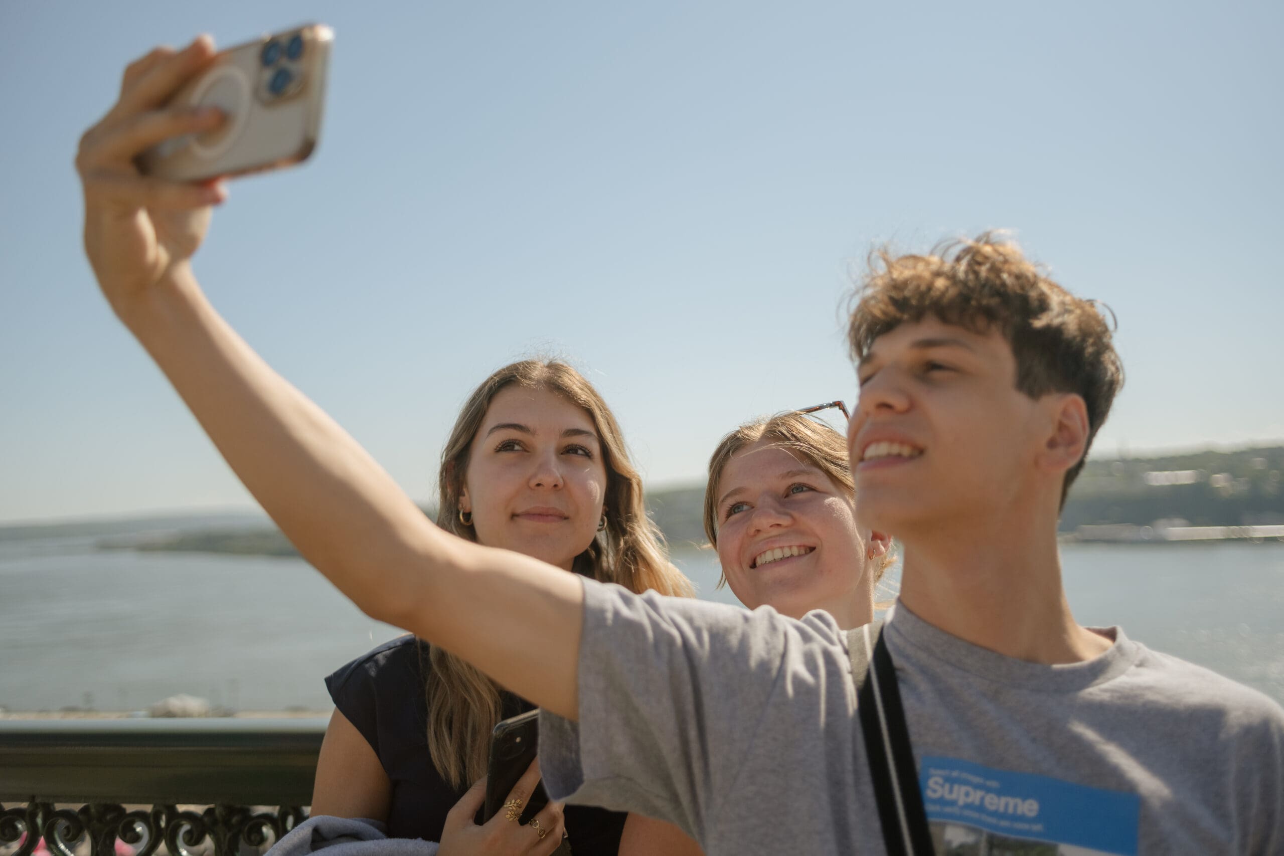 Trois jeunes sourient et prennent la pose pour un selfie en plein air par une journée ensoleillée, avec une rivière et un paysage lointain en arrière-plan.