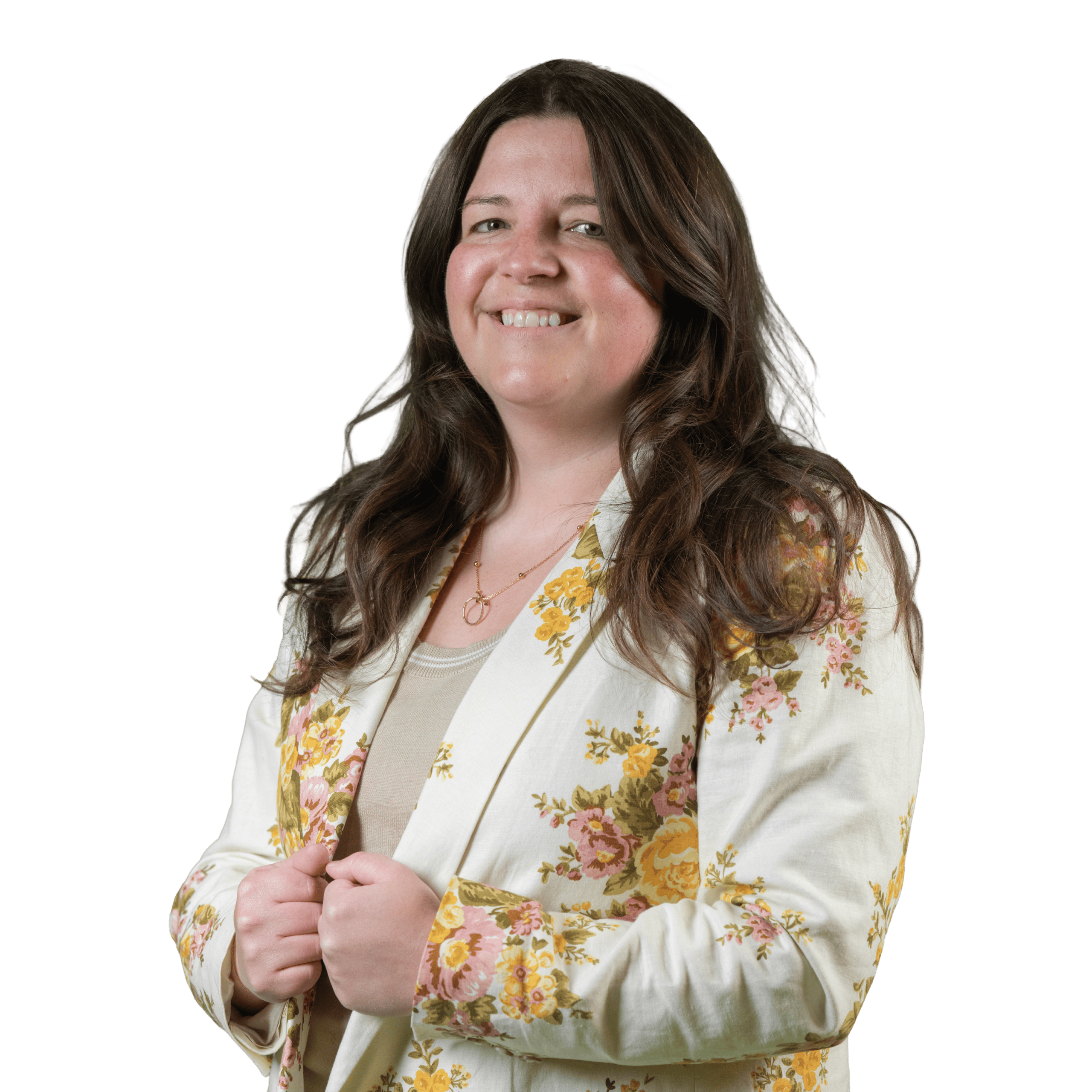 Une femme aux longs cheveux châtains sourit, debout devant un fond blanc. Elle porte une veste de couleur crème à motifs floraux jaunes par-dessus un haut beige.