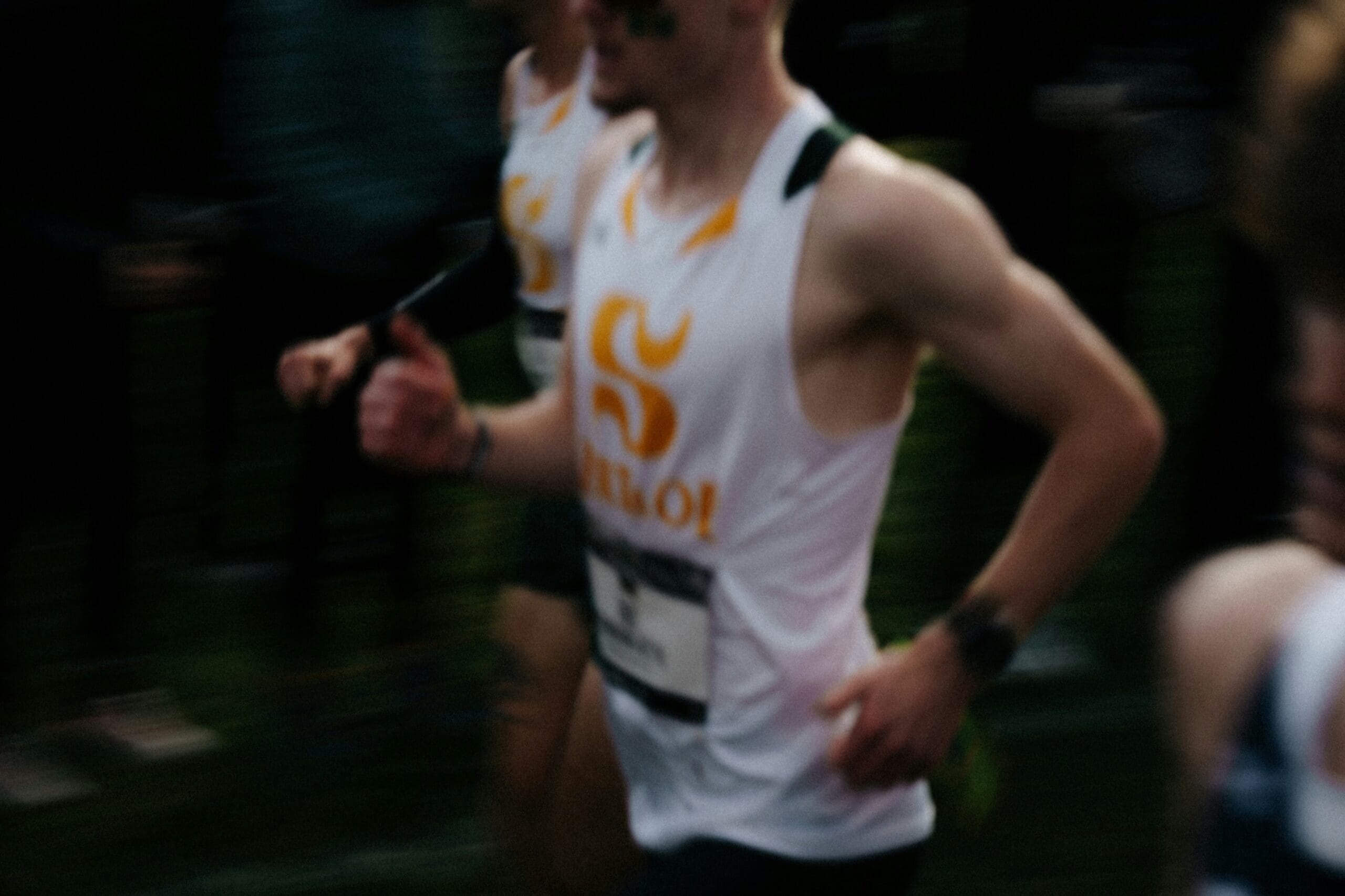 Une image floue représentant un coureur vêtu d'un maillot de sport sans manches blanc et jaune, en pleine foulée lors d'une course, avec d'autres concurrents visibles à l'arrière-plan.