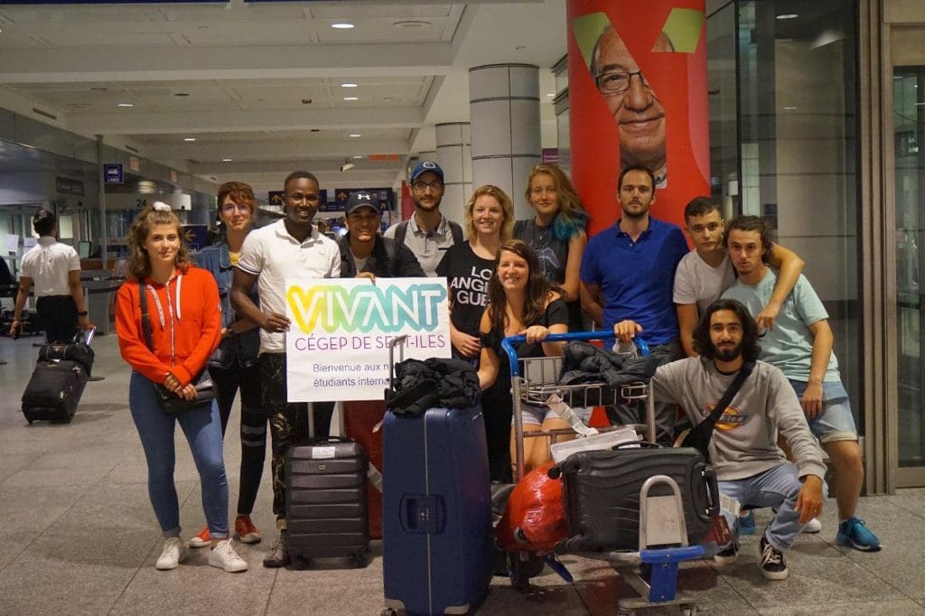 Un groupe de treize personnes, dont certaines tiennent une pancarte sur laquelle on peut lire « VIVANT CÉGEP DE SEPT-ÎLES », se tient debout dans un aéroport, entouré de chariots à bagages et de valises, et sourit à la caméra.