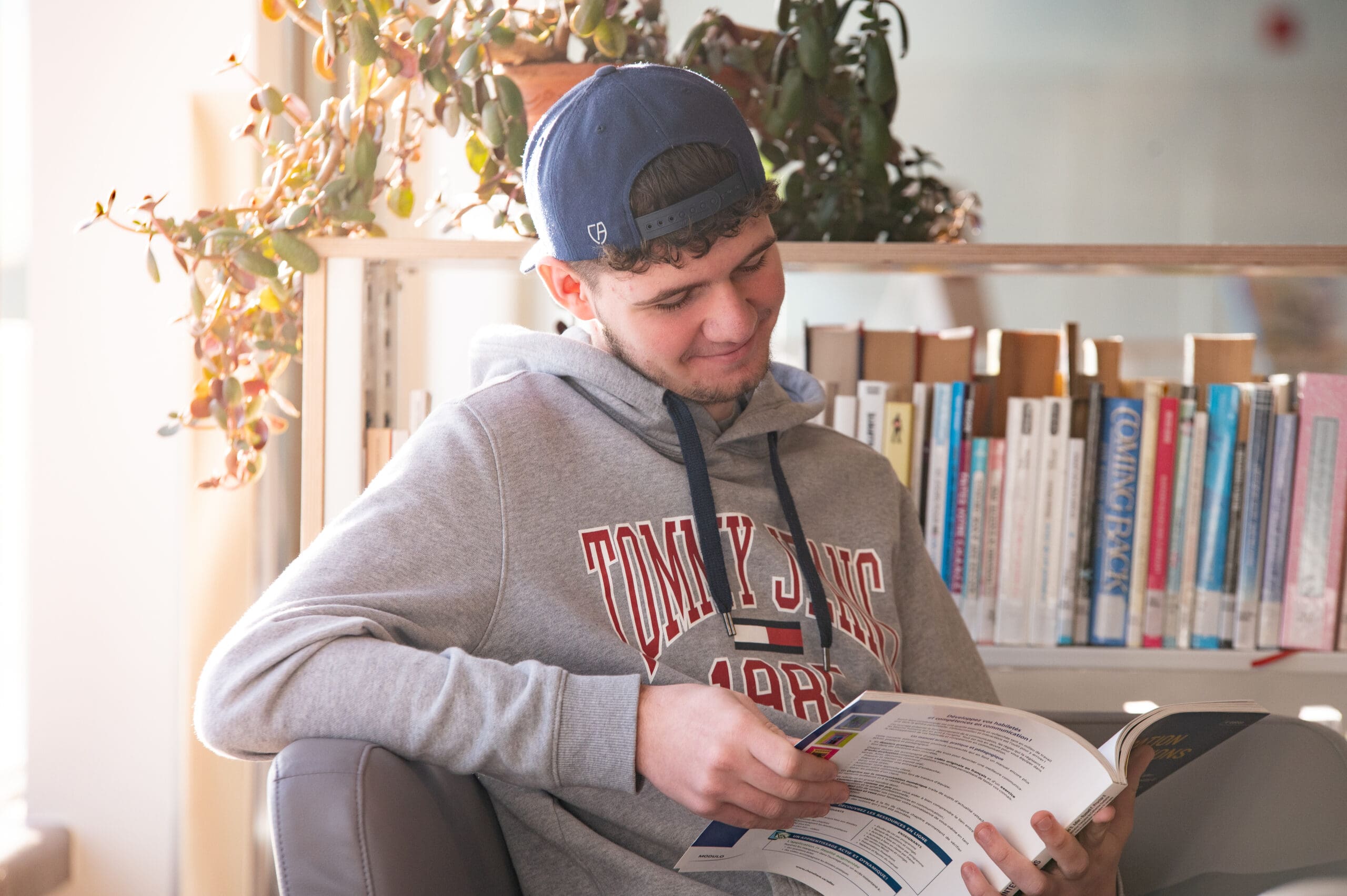 Un jeune homme vêtu d'un sweat à capuche gris et d'une casquette bleue est assis dans une bibliothèque ; il sourit tout en lisant un magazine. On aperçoit des livres et une plante en pot à l'arrière-plan, tandis que la lumière du soleil pénètre par la fenêtre.