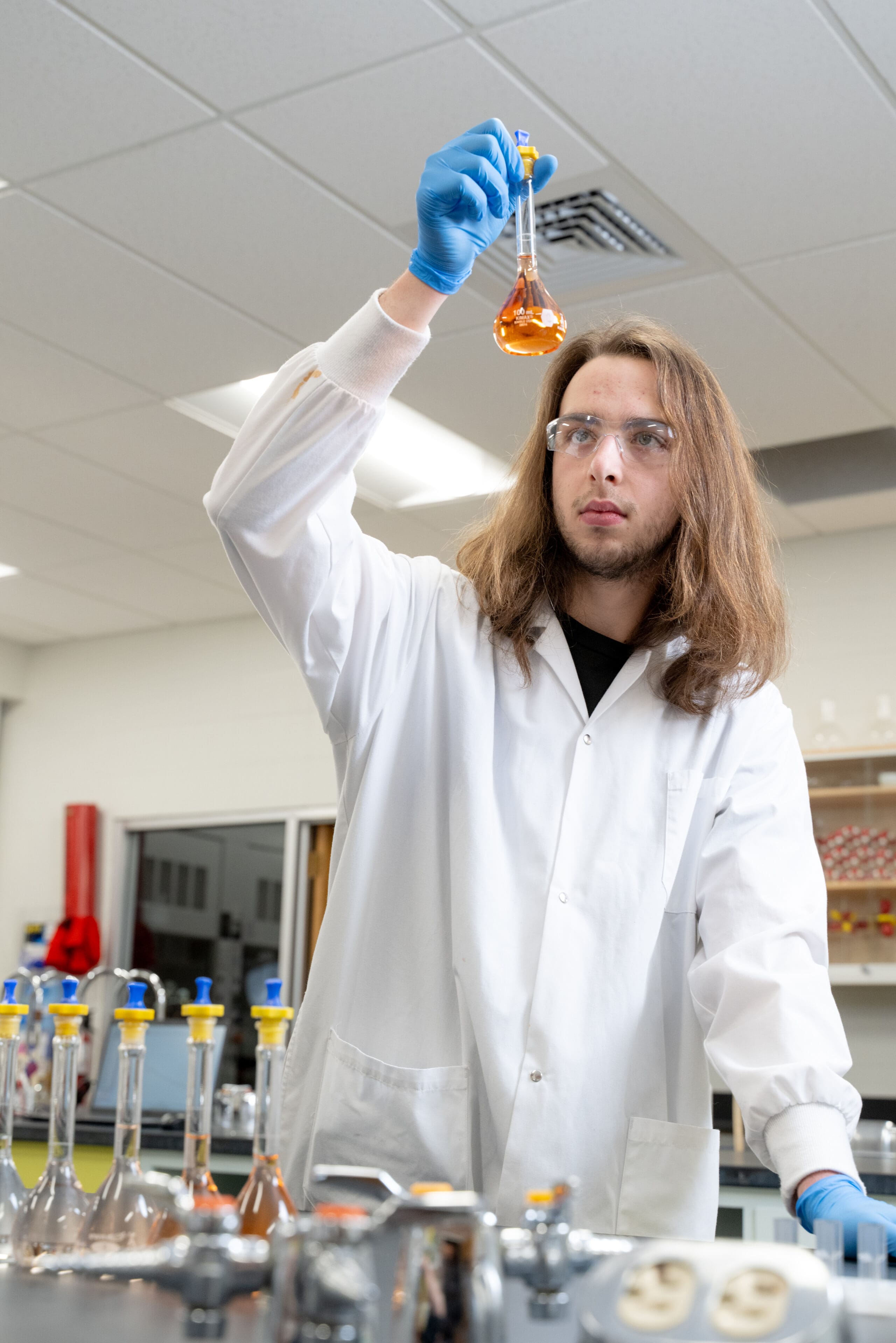 Un scientifique, vêtu d'une blouse de laboratoire et portant des lunettes de protection, tient un ballon contenant un liquide orange et l'examine attentivement. Du matériel de laboratoire et d'autres ballons sont posés sur le plan de travail devant lui.