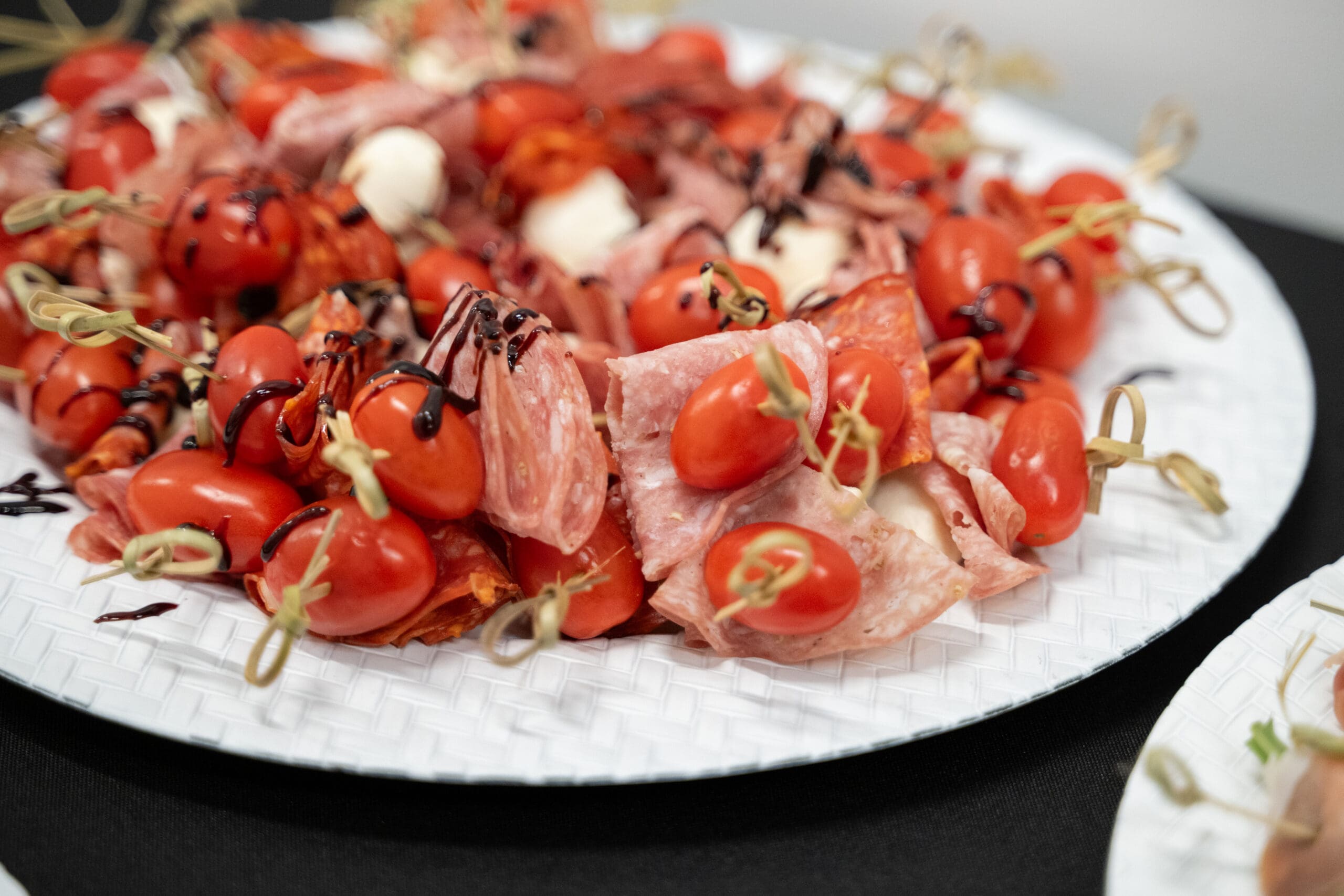 Une assiette blanche présente des brochettes de tomates cerises, de charcuterie, de mozzarella et un filet de glace au vinaigre balsamique, servies en amuse-bouches.
