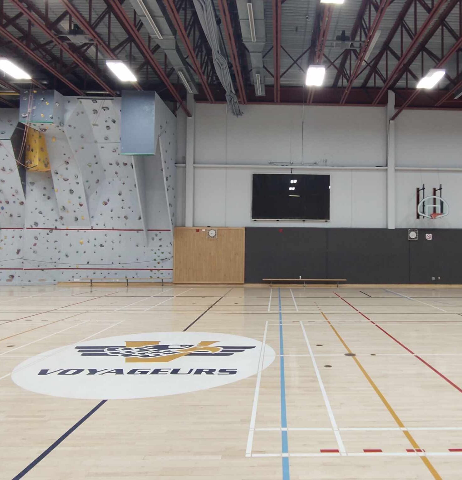 Un vaste gymnase couvert est équipé d'un mur d'escalade, d'un panier de basket et d'un grand emblème représentant le mot « Voyageurs » tracé sur le parquet ciré. On y distingue également des plafonniers et des lignes de terrain peintes.
