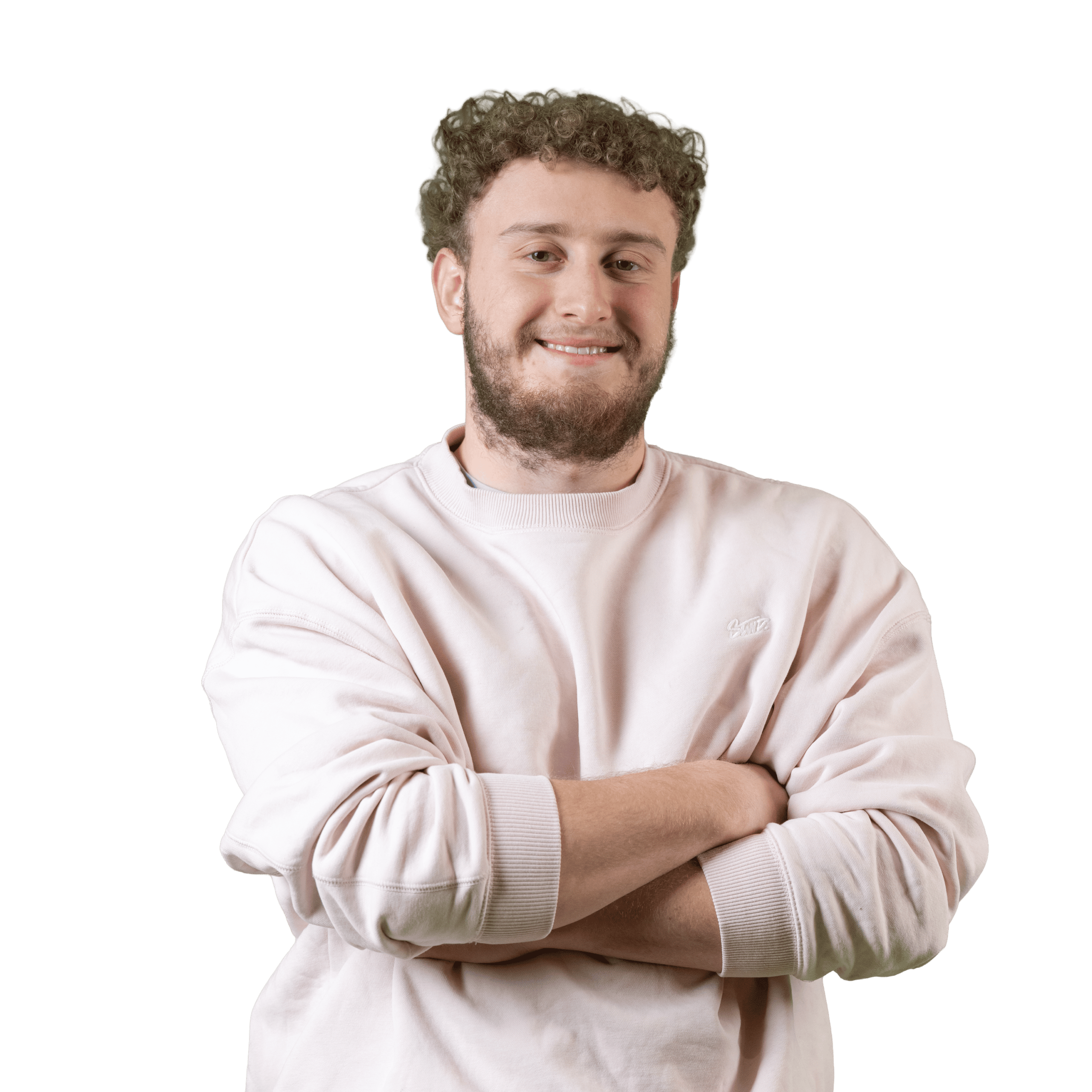Un homme aux cheveux bouclés et à la barbe sourit, les bras croisés, vêtu d'un sweat-shirt rose pâle sur un fond blanc uni.