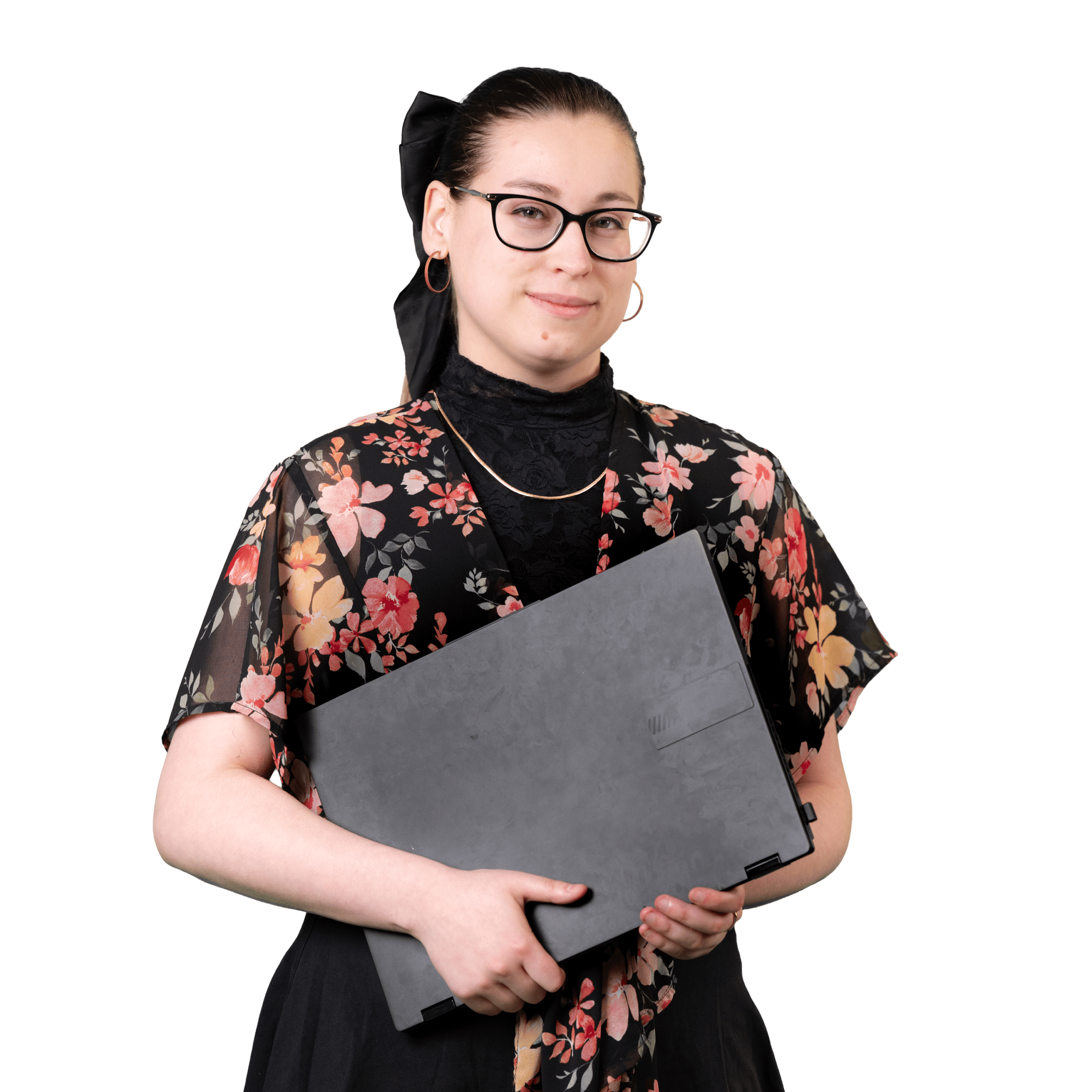 Femme portant des lunettes et un haut à motifs floraux, tenant devant elle un ordinateur portable noir fermé, debout devant un fond blanc uni.
