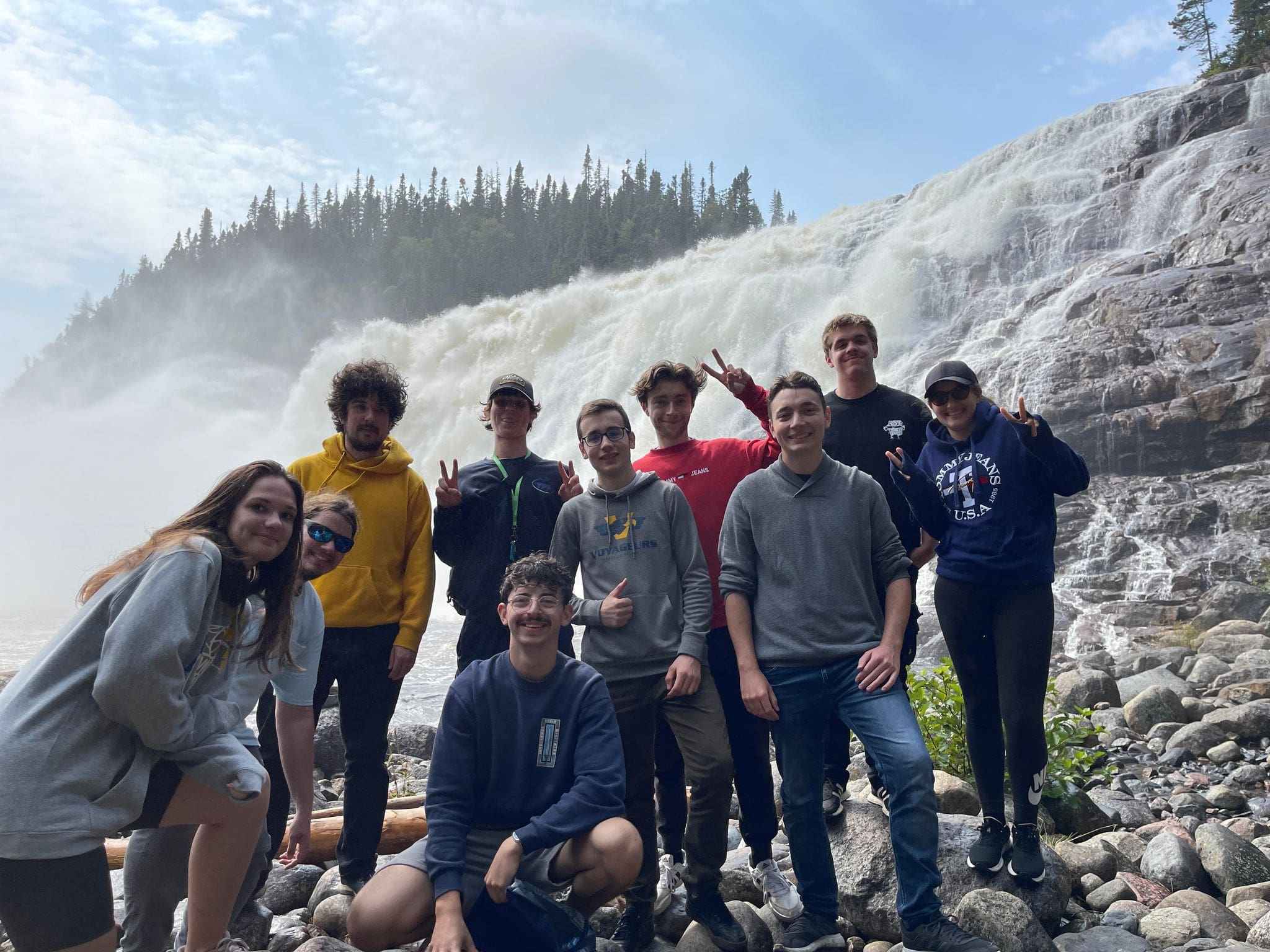 Onze personnes prennent la pose et sourient sur un sol rocailleux, devant une grande cascade enveloppée de brume et des pins, sous un ciel partiellement nuageux.