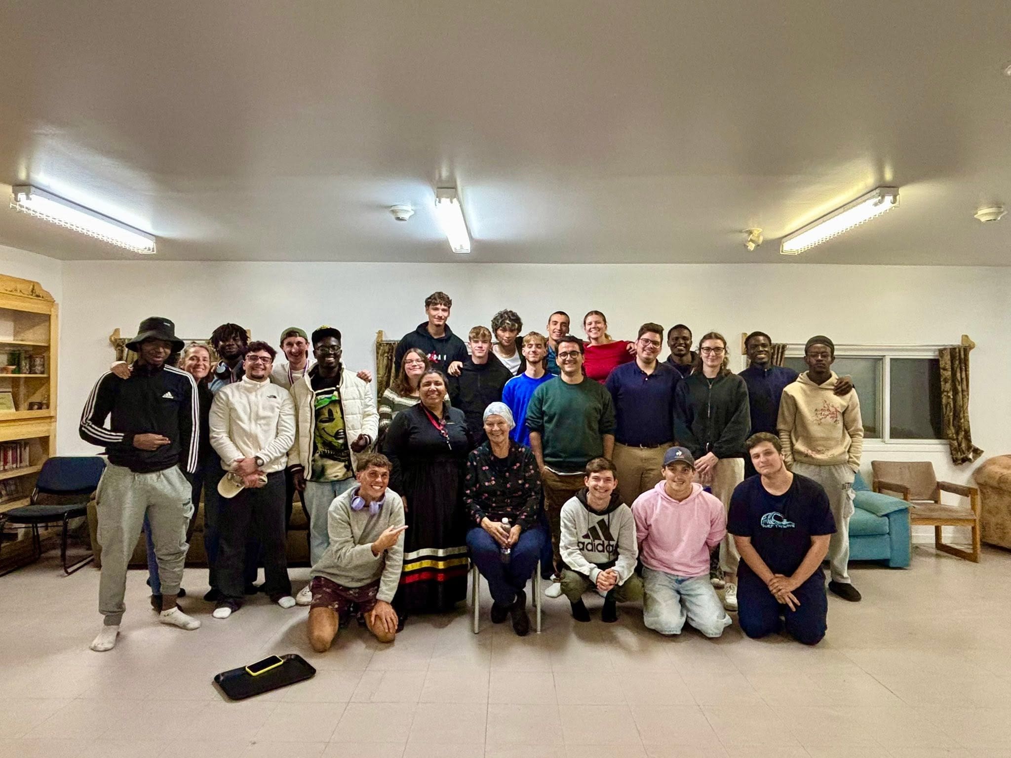 Un groupe d'environ 28 personnes, composé principalement de jeunes adultes et de quelques personnes plus âgées, pose ensemble à l'intérieur d'une pièce très lumineuse aux murs blancs, meublée de canapés et de bibliothèques. Tout le monde sourit à l'appareil photo.